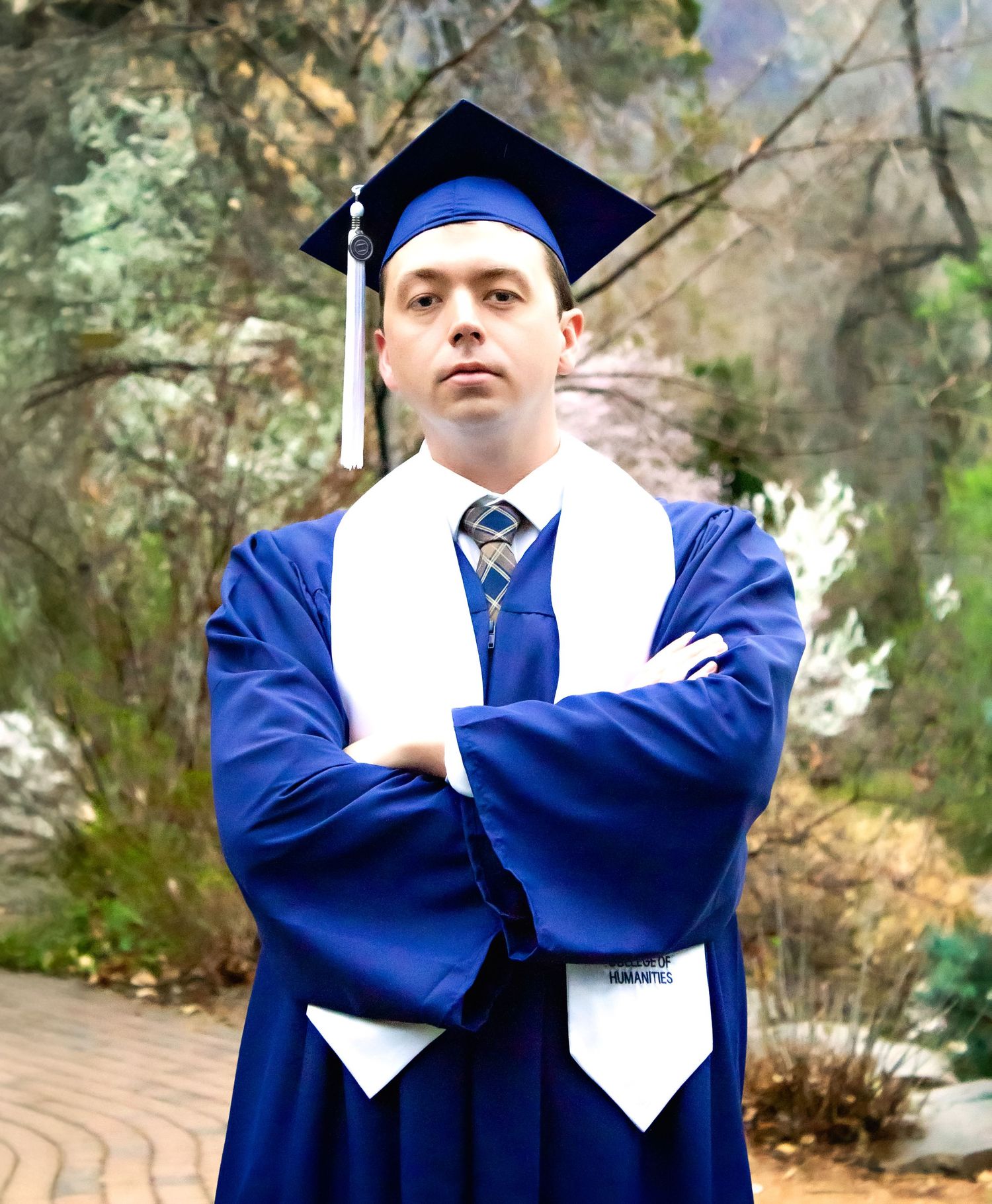 Graduate in blue cap and gown stands with crossed arms against natural outdoor setting.