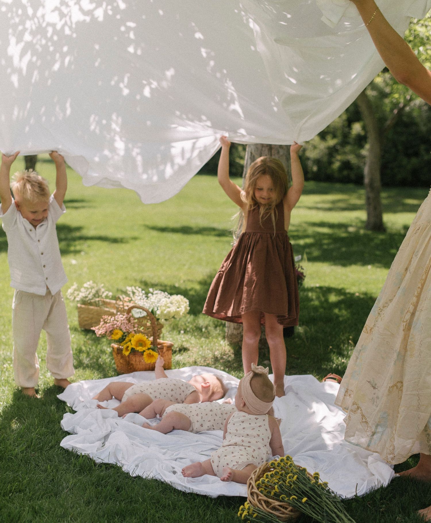 Children playing under a white canopy on a blanket in a sunny garden setting.