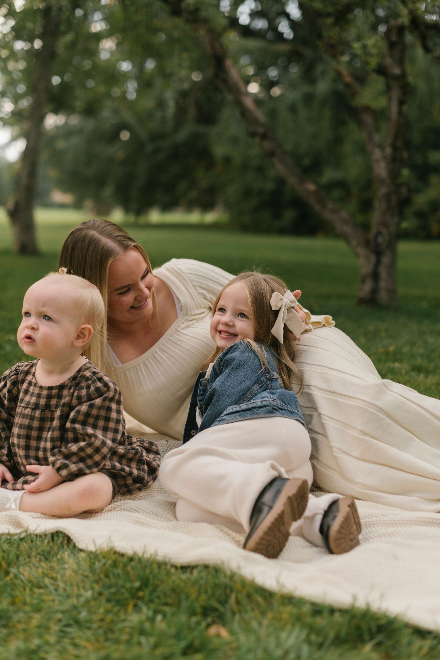 Family relaxing together on white blanket in a park setting.