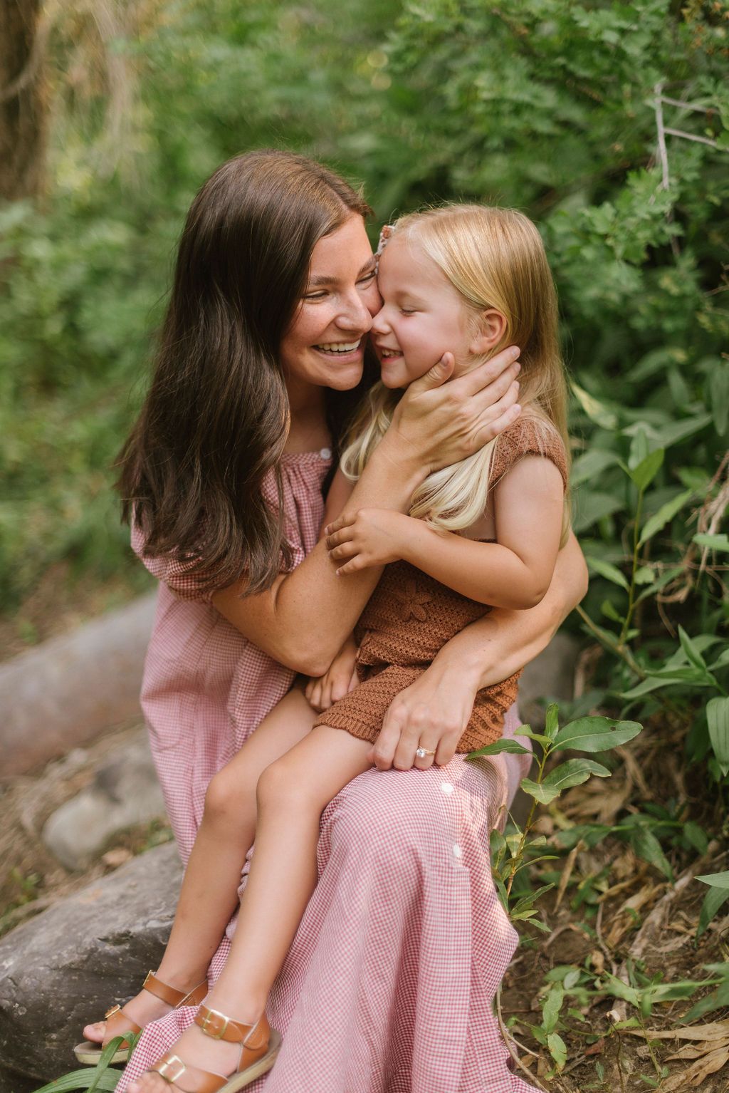 A tender moment between parent and child sitting outdoors in pink dresses against a natural green background.