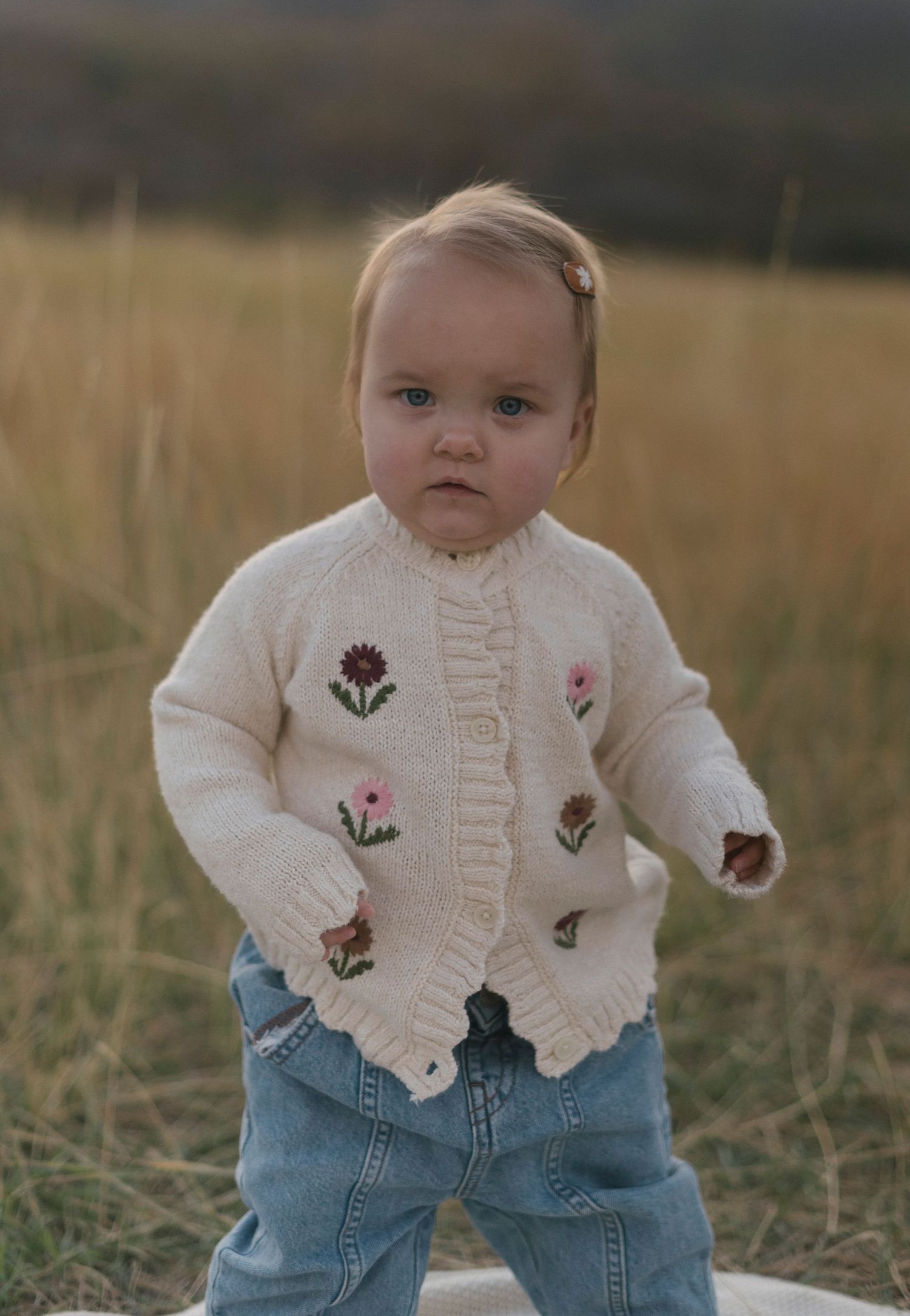 A toddler wearing a floral cardigan and jeans stands in a golden field.