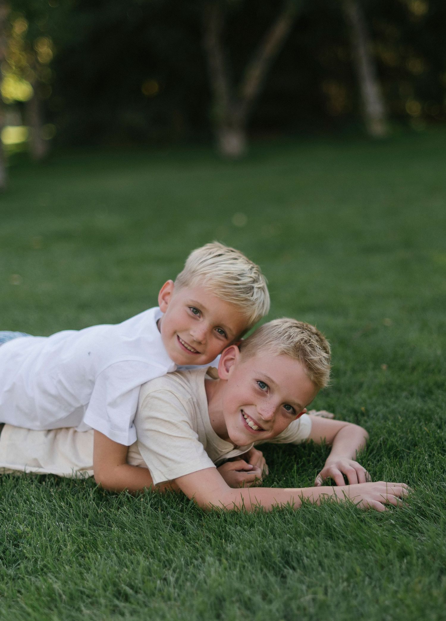 Two children in white shirts lying on green grass while playing together outdoors.