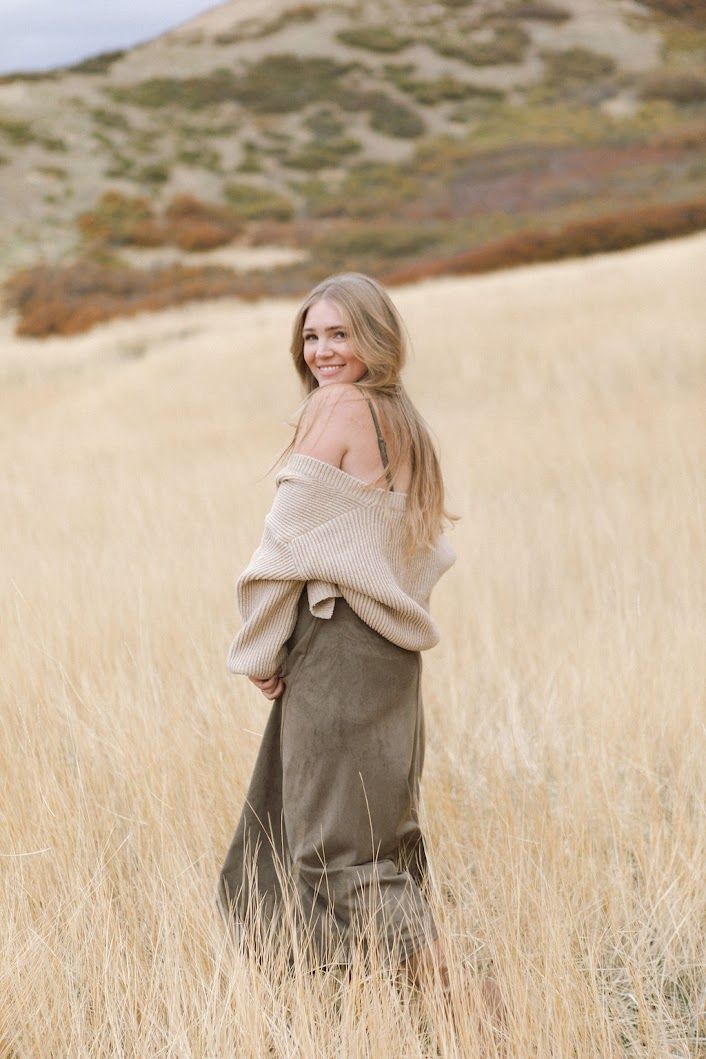 A person twirling in an olive dress against a backdrop of golden grass and rolling hills.