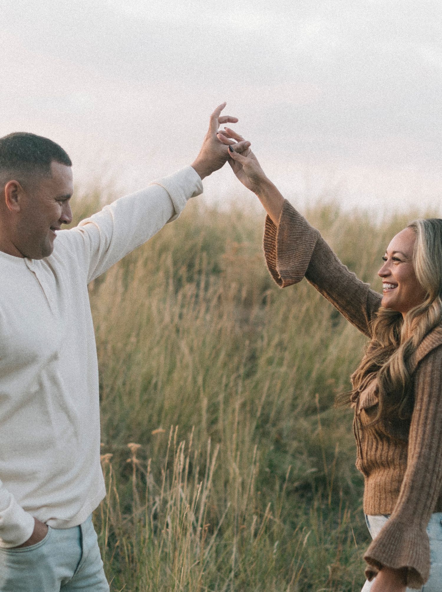 A couple shares a joyful dancing moment together in a field of tall grass at sunset.