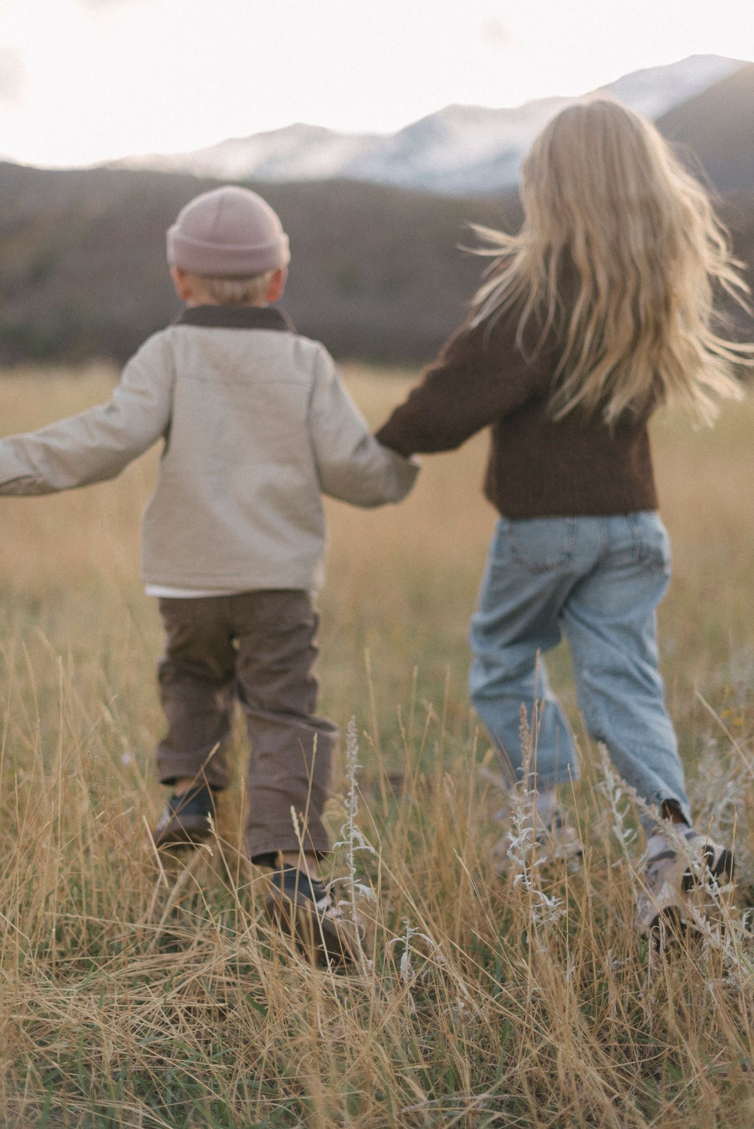 Two children walking together through tall grass at sunset with mountains in background.