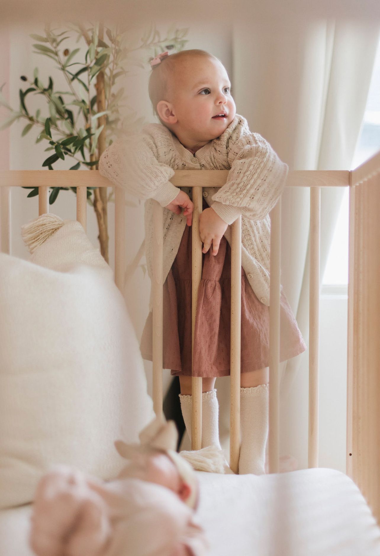 Young child in a cream sweater peering over wooden crib railing in a bright nursery.