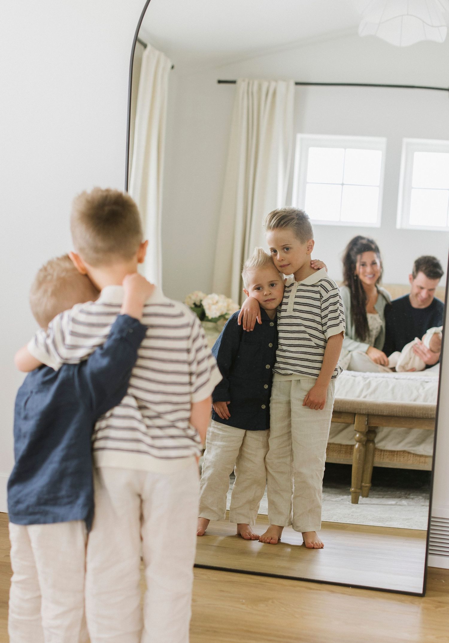 Children looking at their reflection in a bedroom mirror with family visible in background.