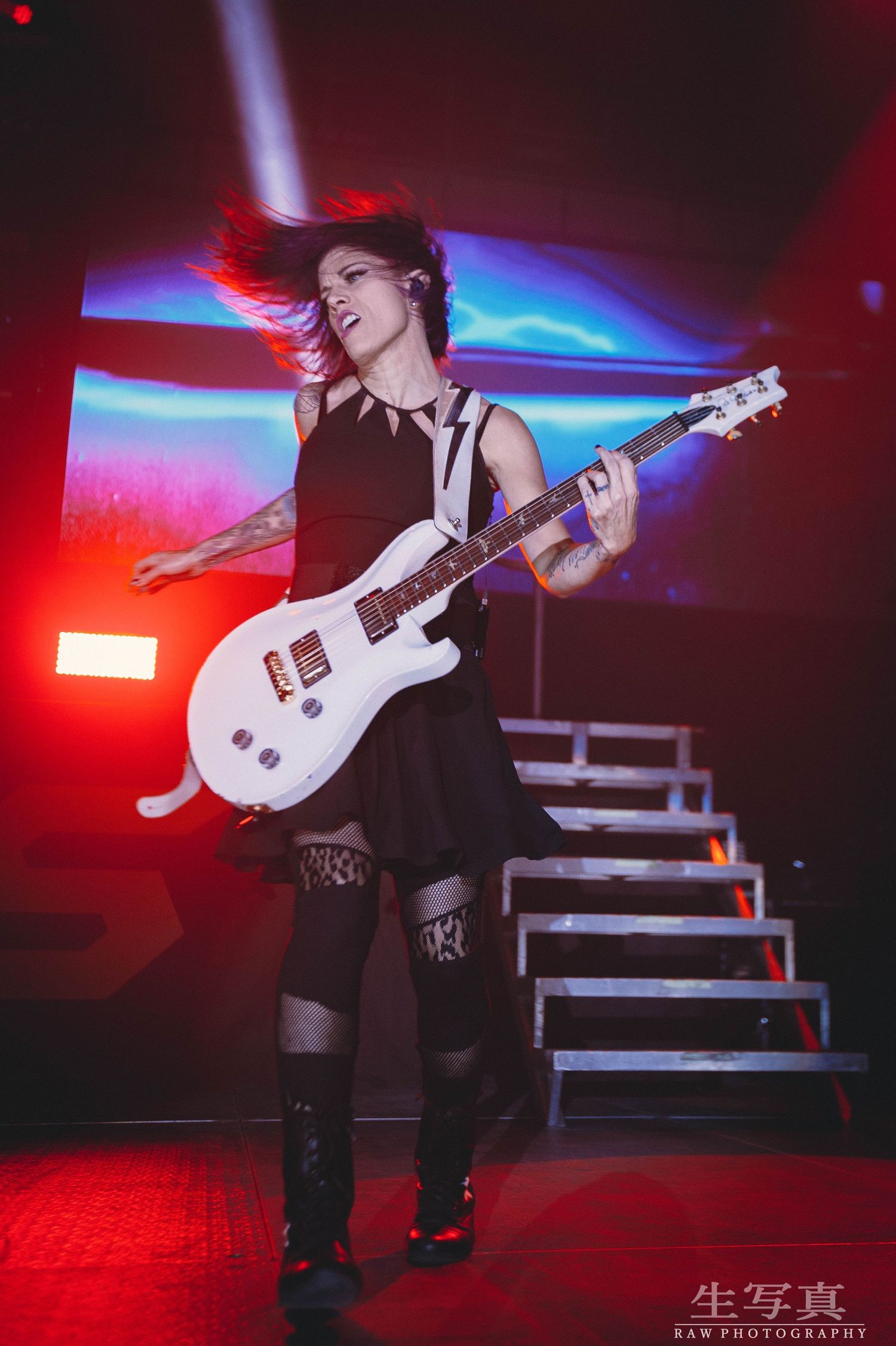 Guitarist with white electric guitar performing energetically under red stage lights.
