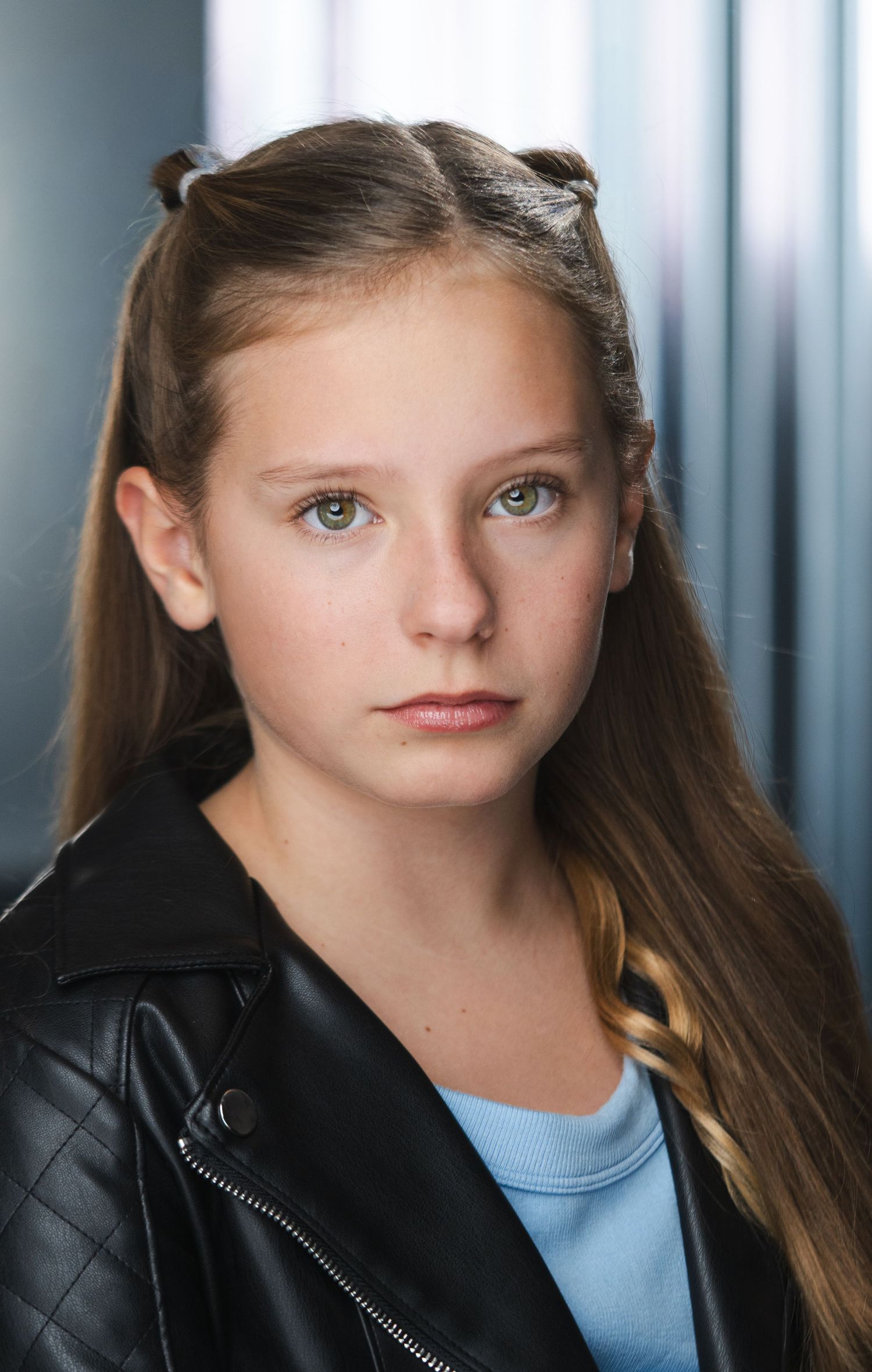 Young person with long brown hair poses in black jacket against blurred background.