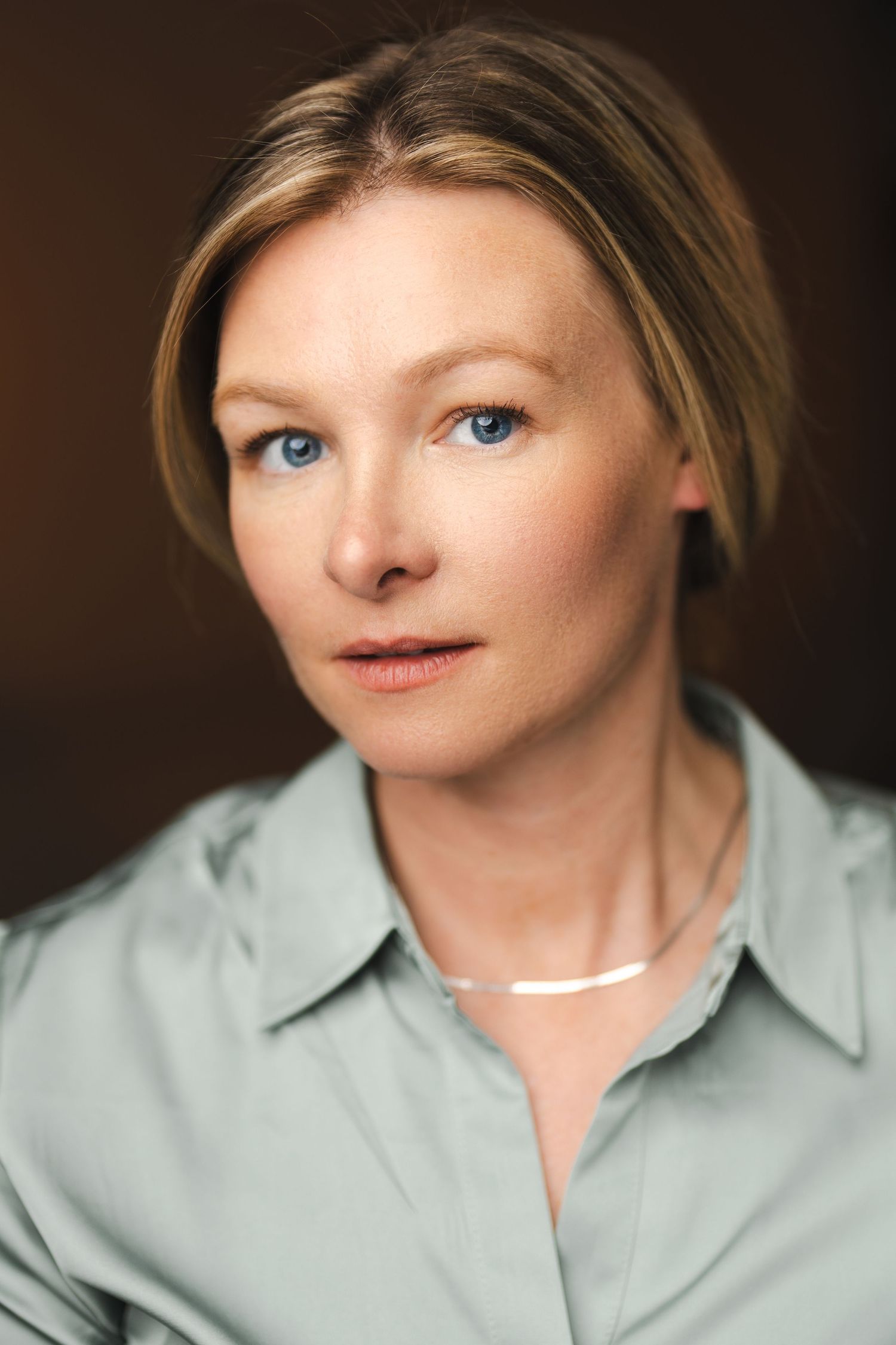 Portrait of person in sage green blouse with delicate necklace against dark background.