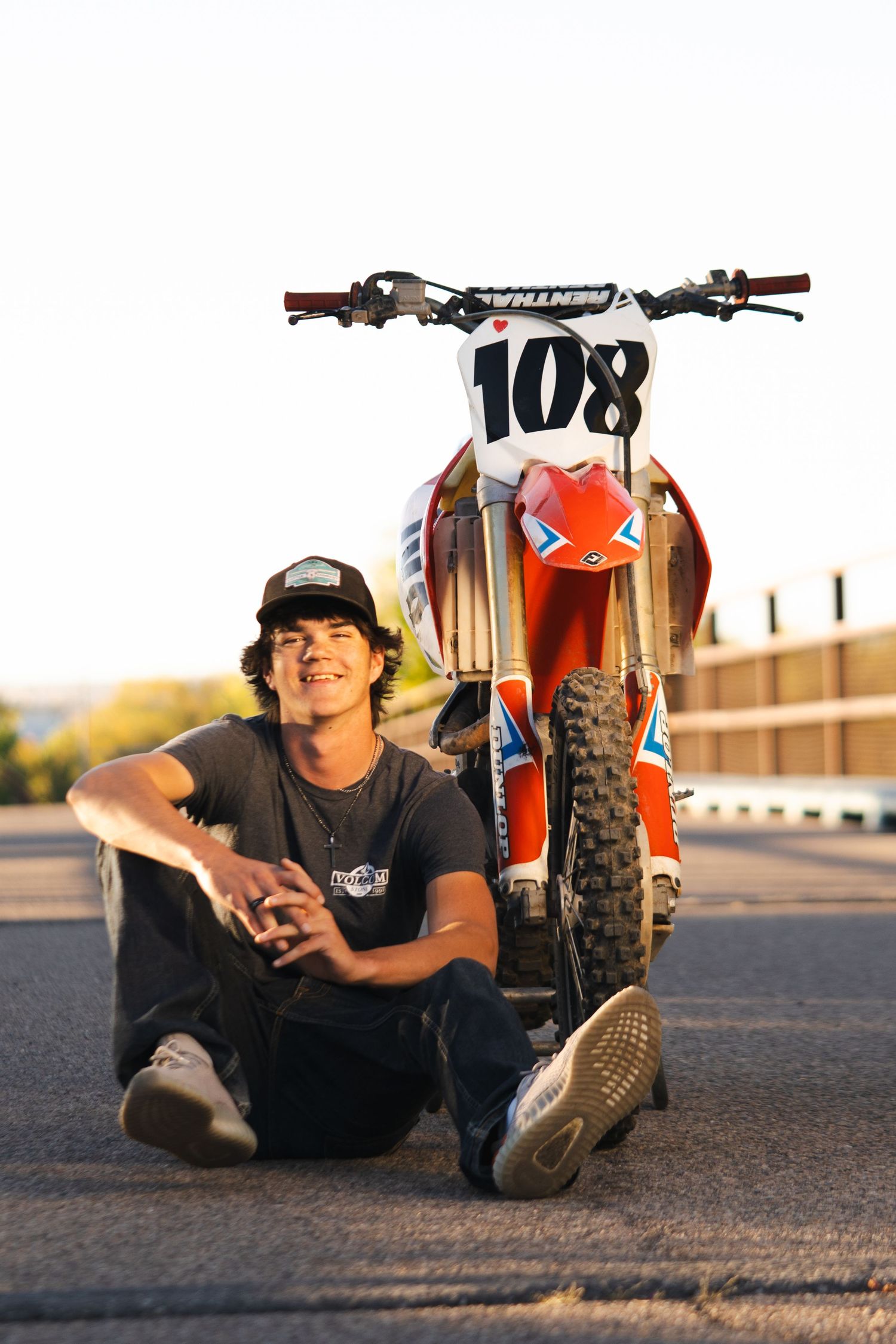 Casual portrait with orange dirt bike on asphalt during golden hour.