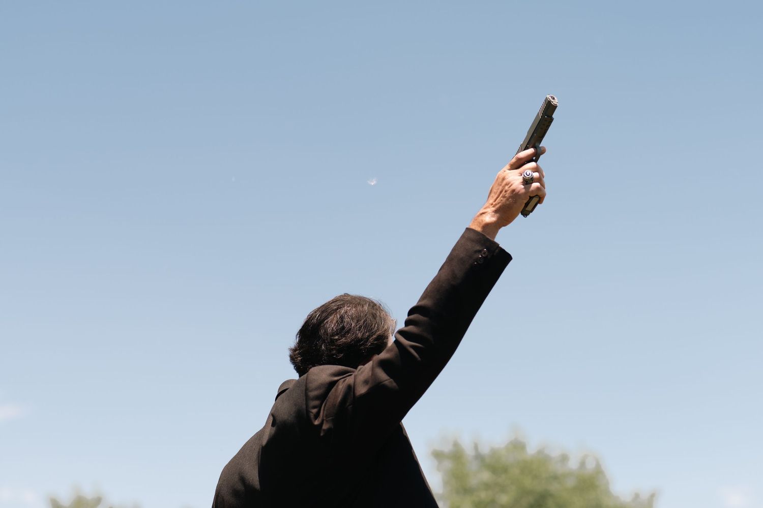 Person in dark jacket firing gun into air against blue sky background.