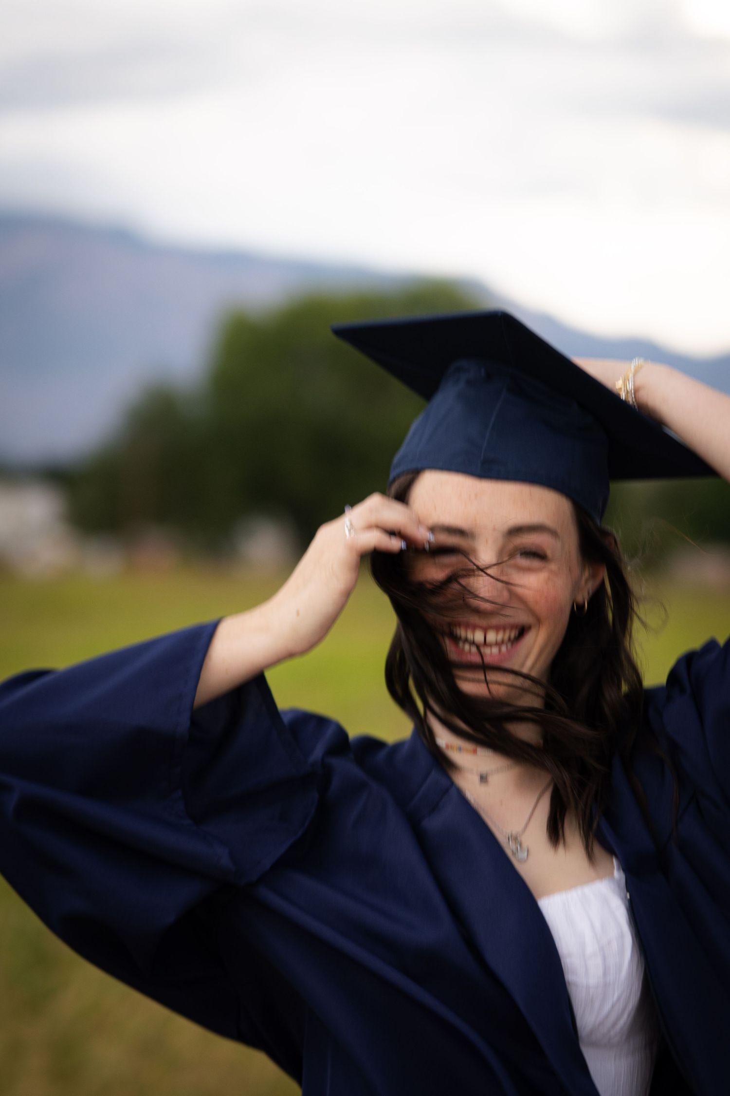 Graduate in navy cap and gown laughs while adjusting mortarboard outdoors.