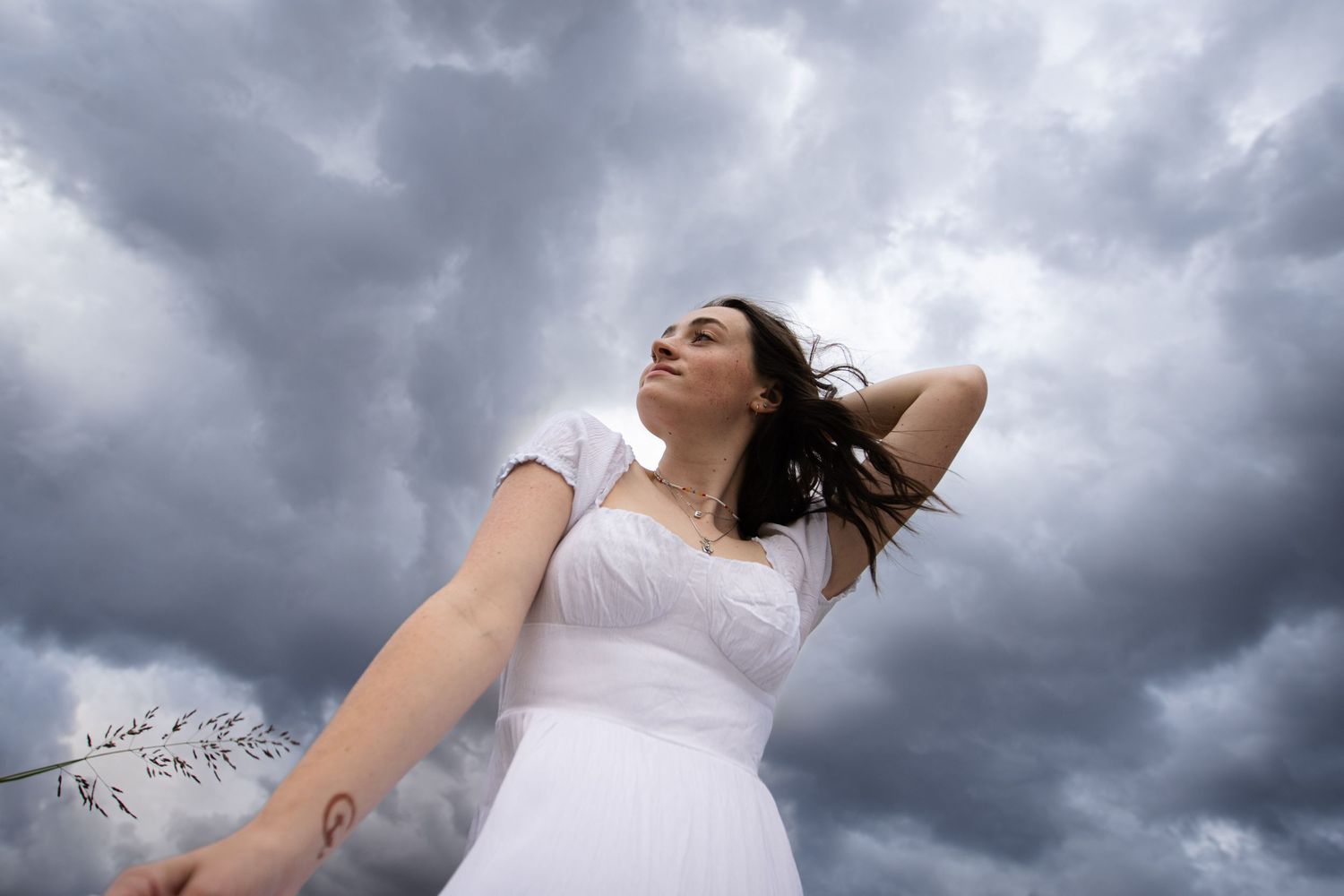 A woman in a white dress poses dramatically against stormy clouds with her hair flowing in the wind.