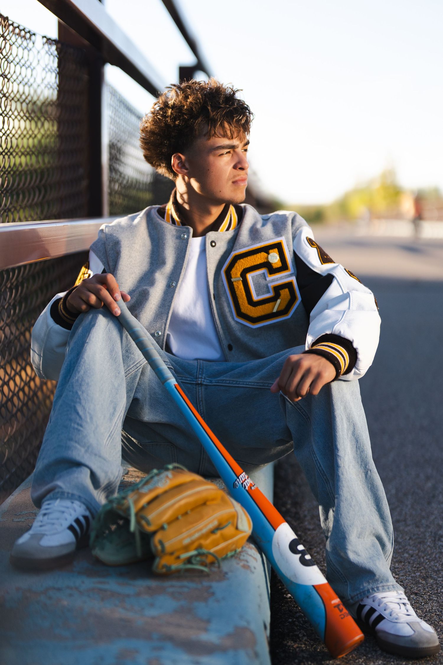 Person in varsity jacket sitting with baseball glove and bat on concrete surface.