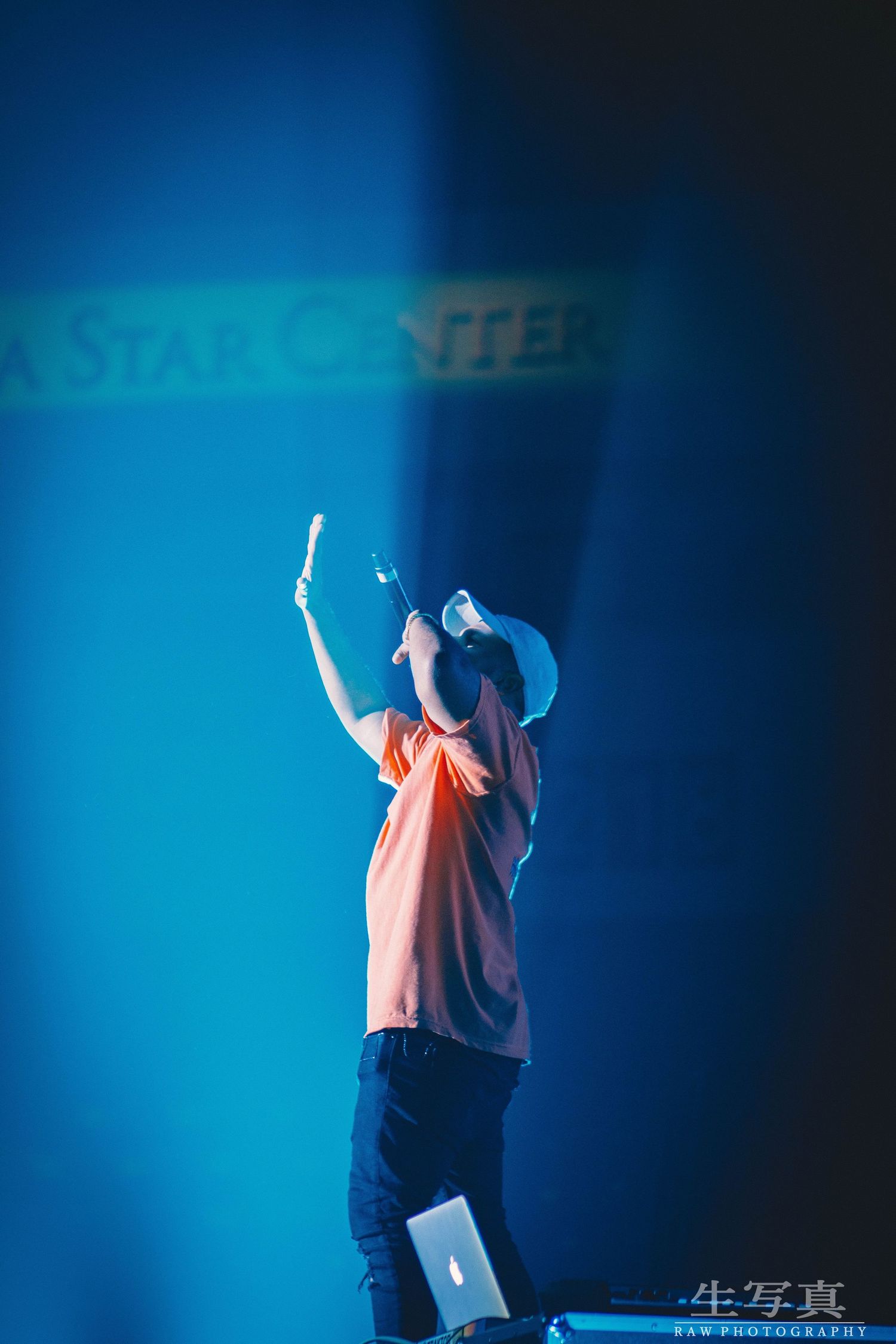 Performer in orange shirt silhouetted against blue backdrop raising arms during concert performance.