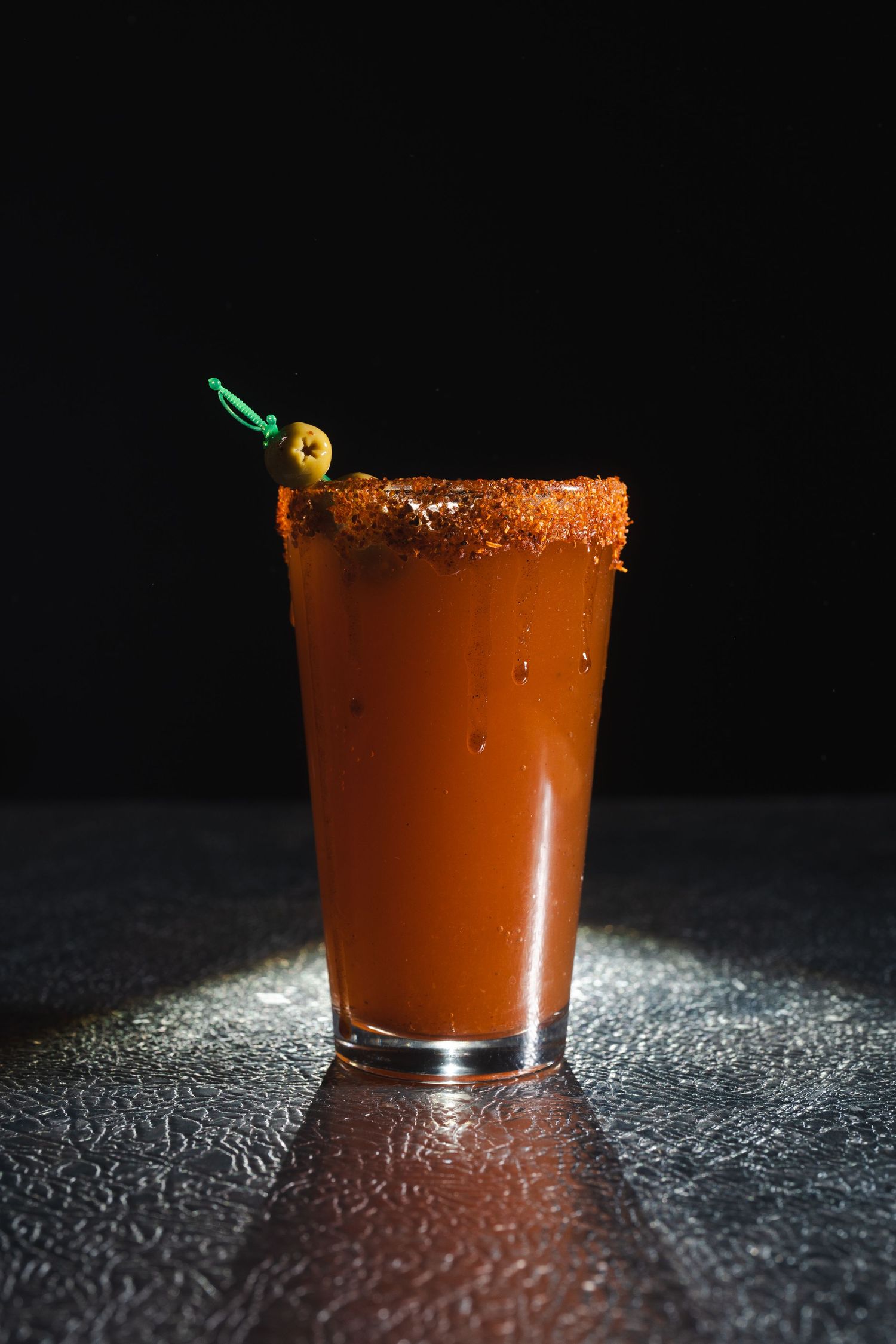 Spicy red cocktail with salted rim and olive garnish in dramatic lighting.