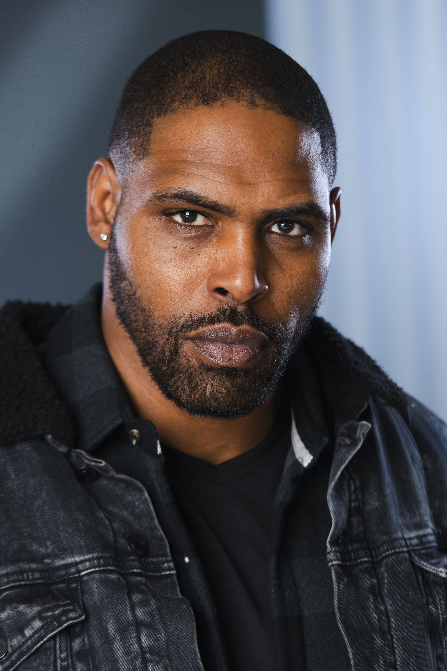 Portrait of a person in a black denim jacket with intense gaze against a blue-tinted background.