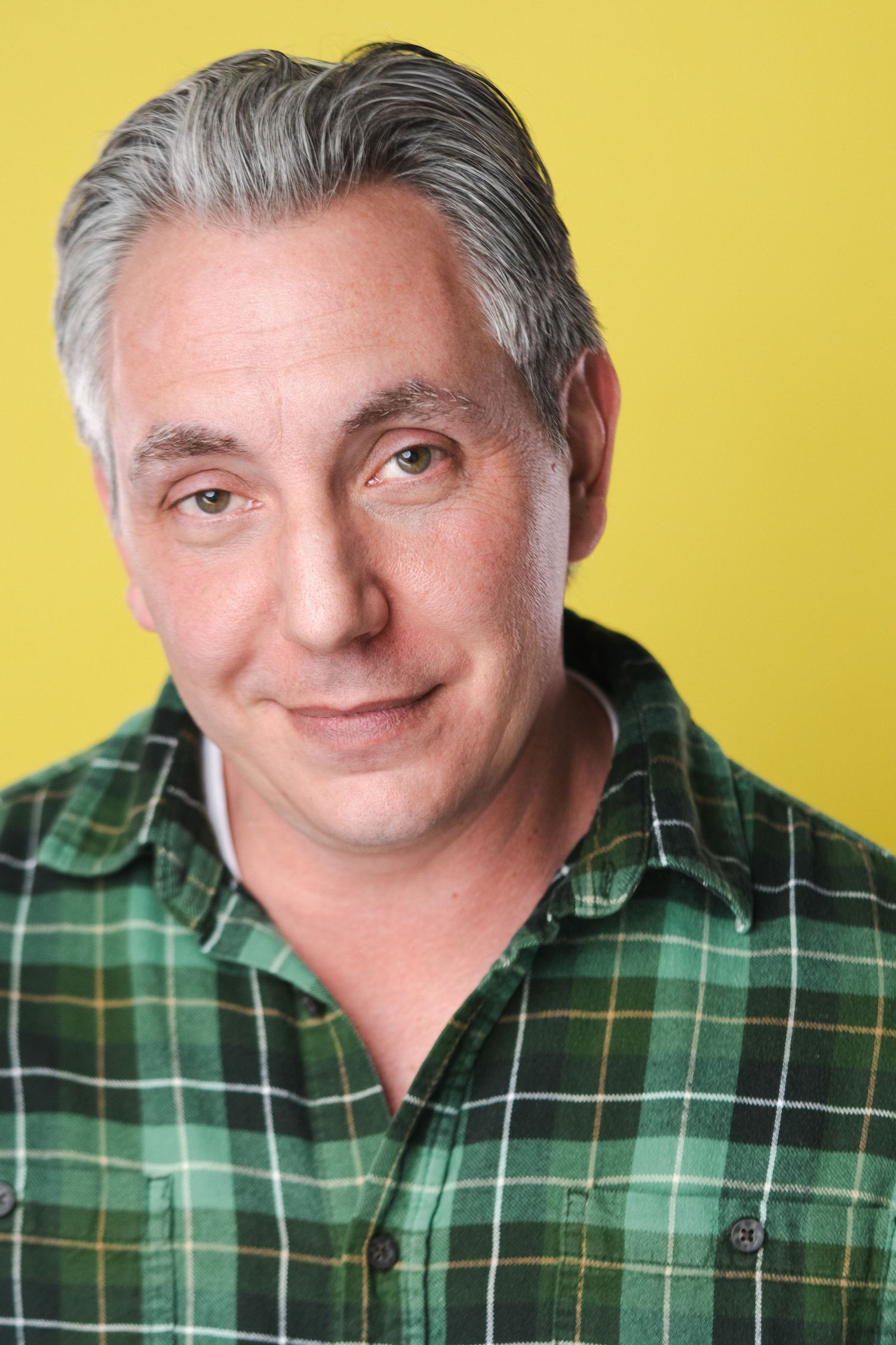 Professional headshot of a person in green plaid shirt smiling warmly against yellow background.