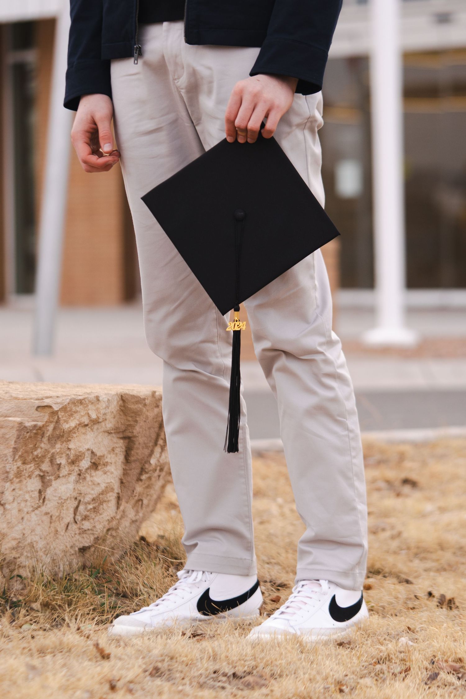 A close-up of legs wearing white Nike sneakers while holding a graduation cap.