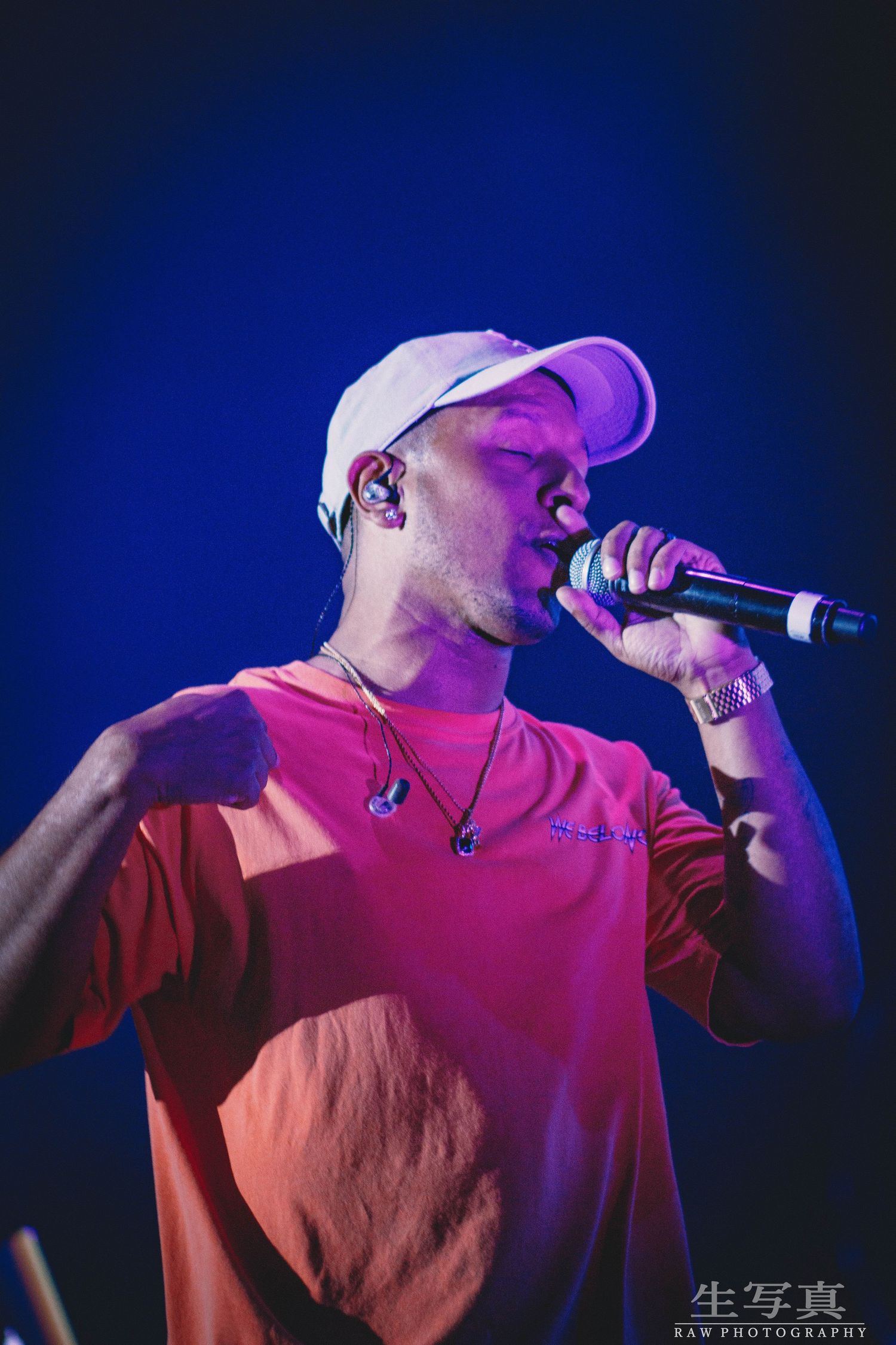 Concert performer in pink shirt and white cap singing emotively under purple and blue stage lights.