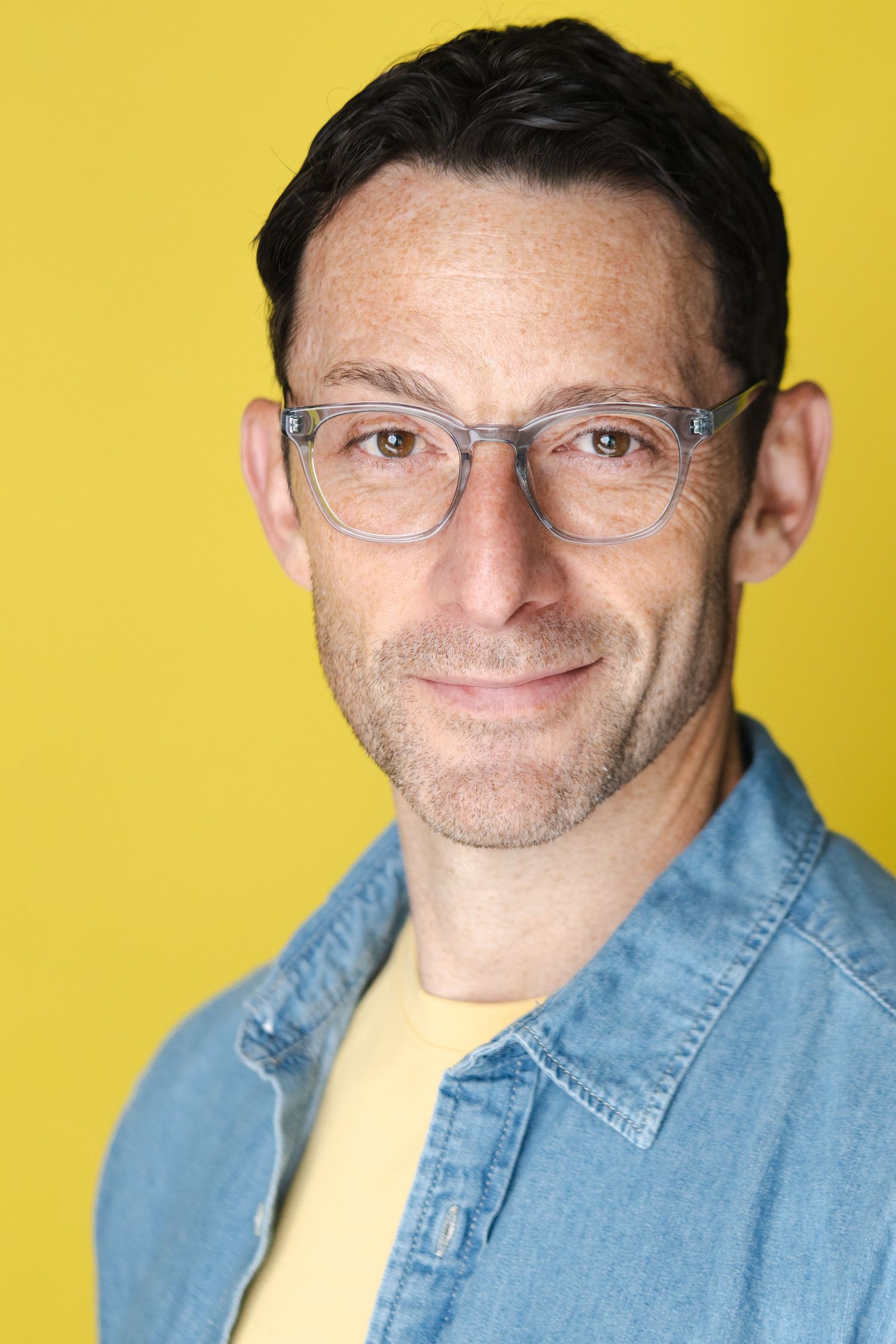 Professional headshot of a person wearing glasses and denim jacket against a bright yellow background.