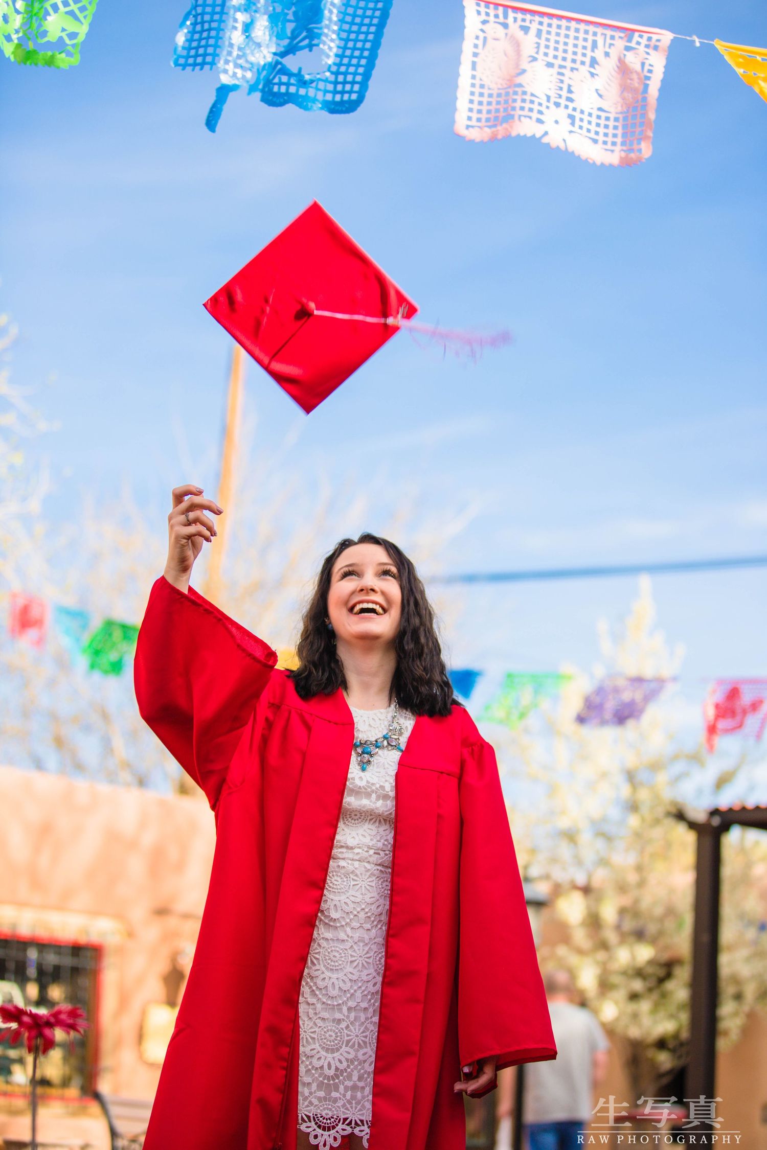 New Mexico Photographer - Graduation Graduate in red gown joyfully holds cap up under festive papel picado decorations.