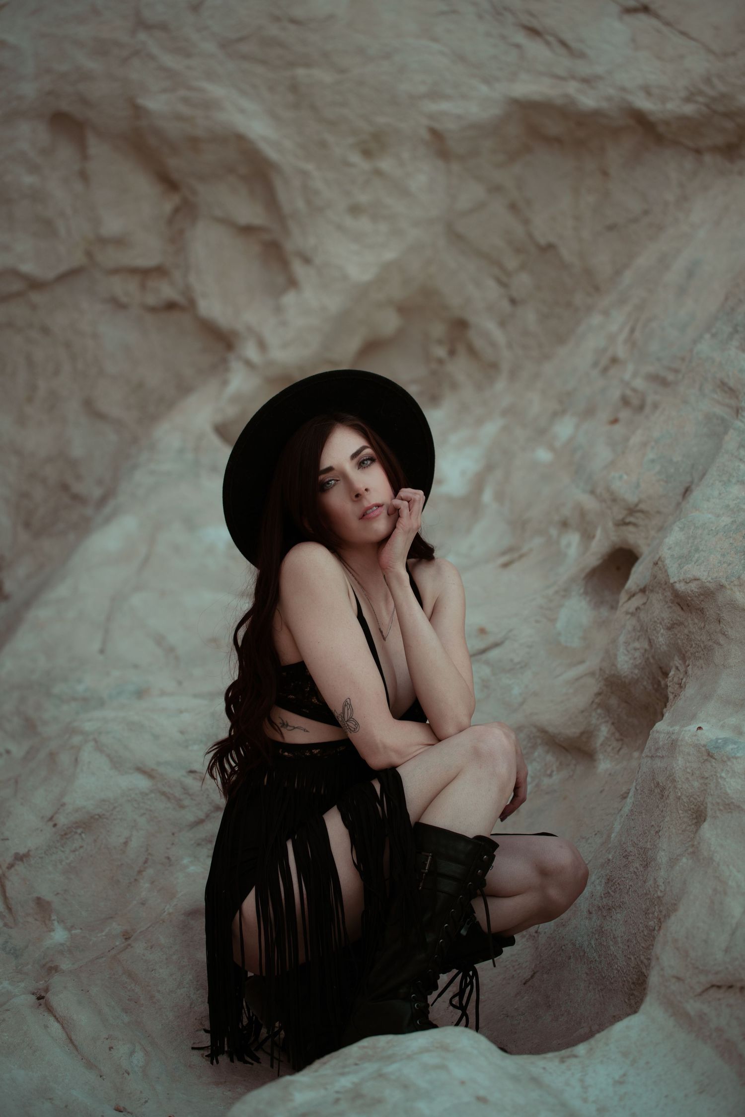 Artistic portrait of a person in black attire and hat posing against textured rock formation.