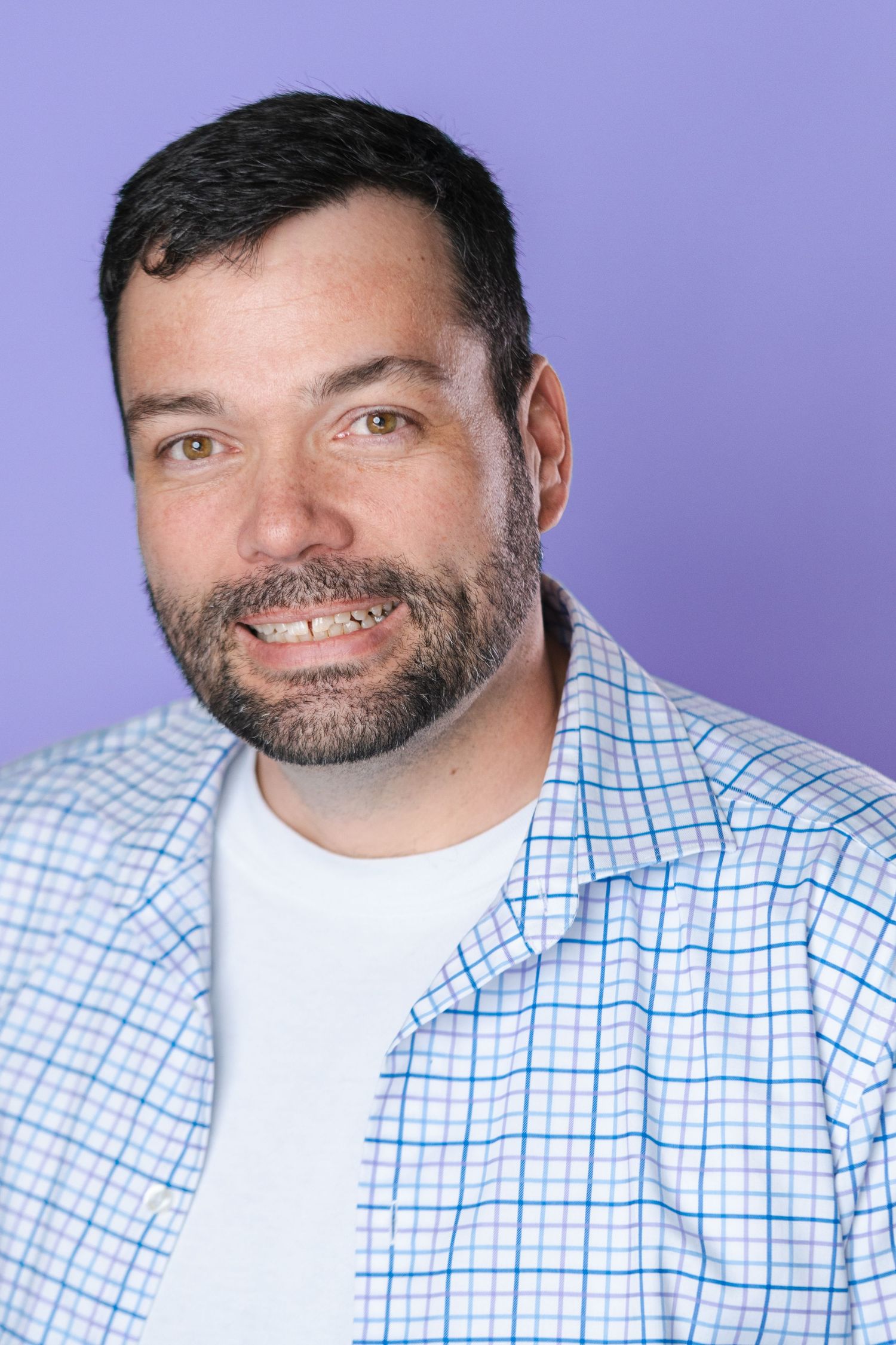 Professional headshot of a person in a light blue checkered shirt smiling warmly against a purple background.