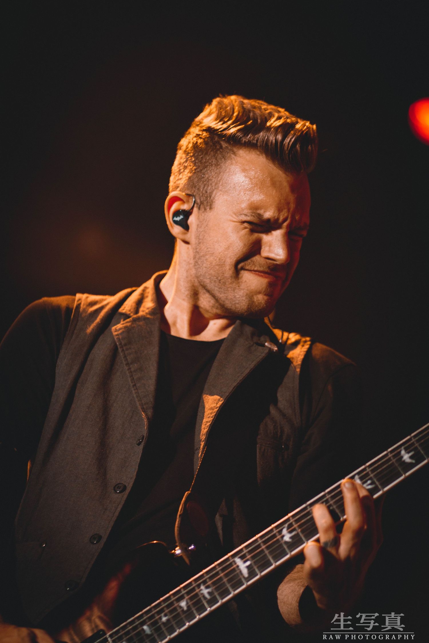 Musician in dark jacket playing electric guitar on dimly lit stage.