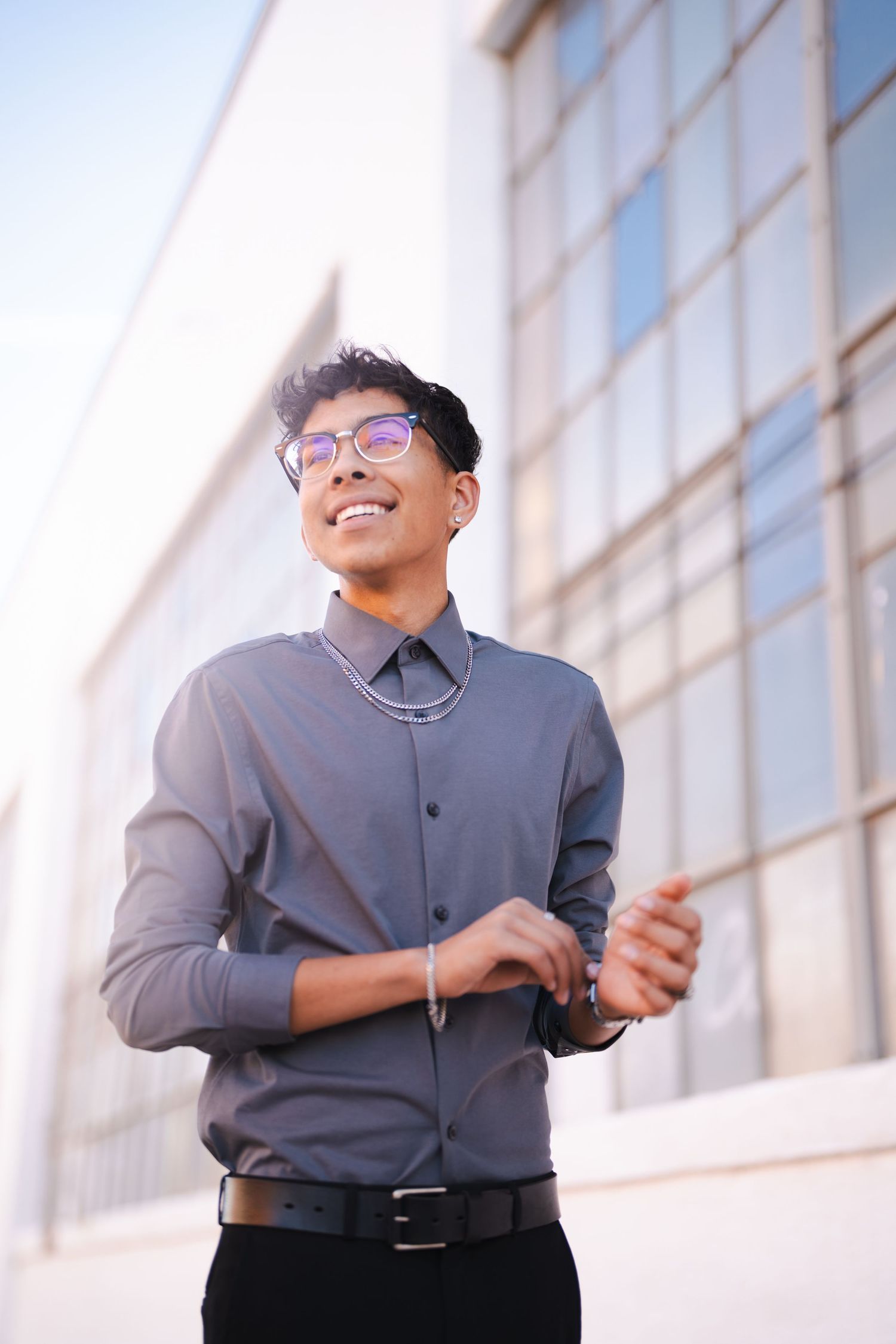 Person in grey dress shirt and glasses standing by large windows in natural light.