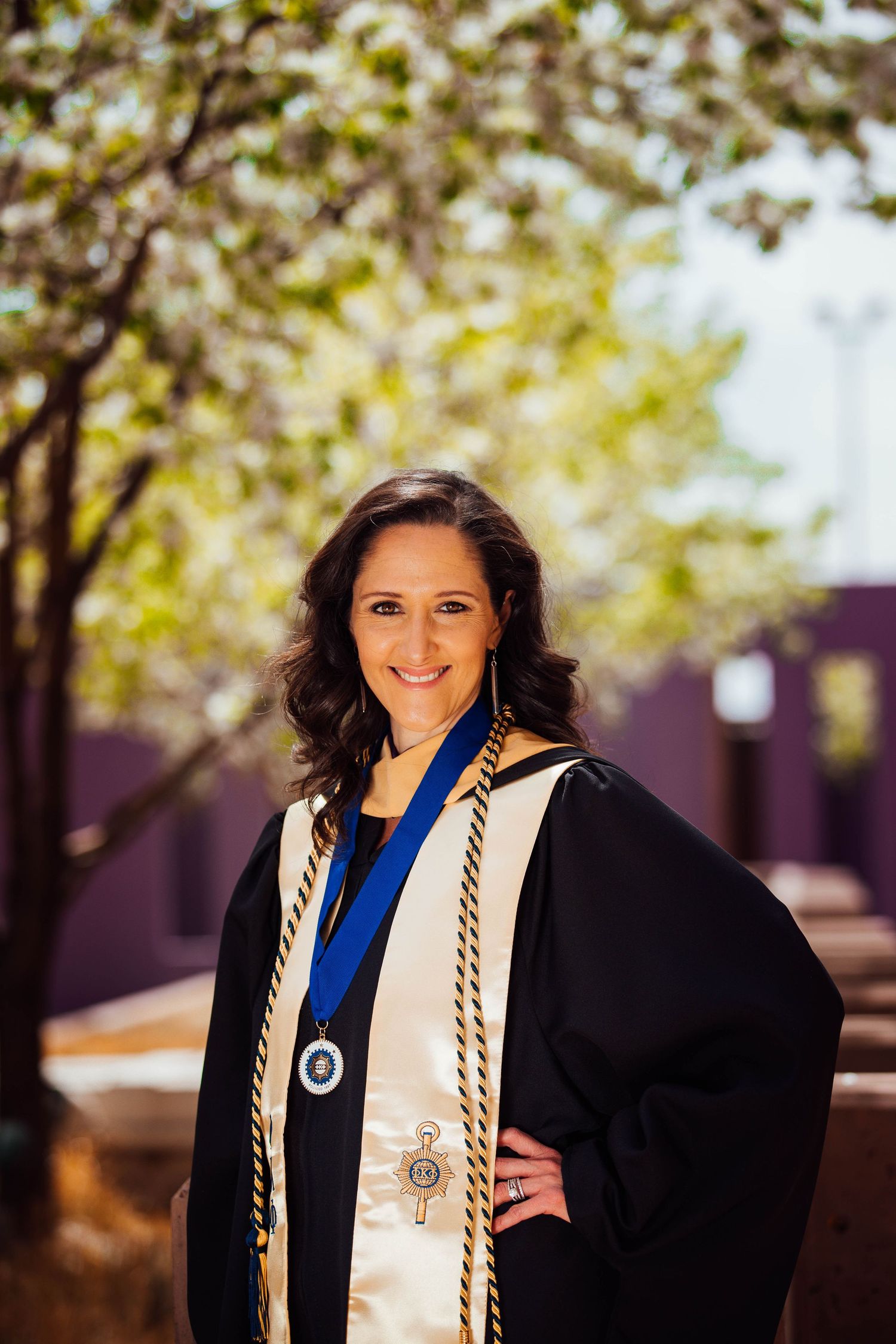 Graduate in black academic regalia with blue stole and white cords poses outdoors under blooming spring trees.