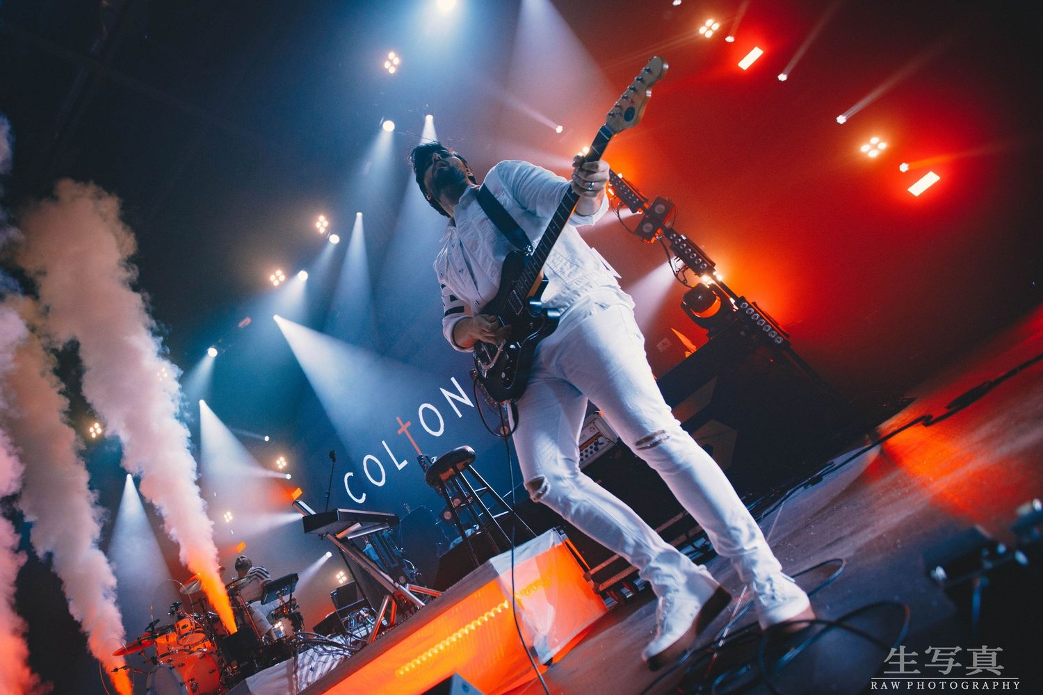 Dynamic concert performer in white outfit leaping on stage with smoke effects and colorful lighting.