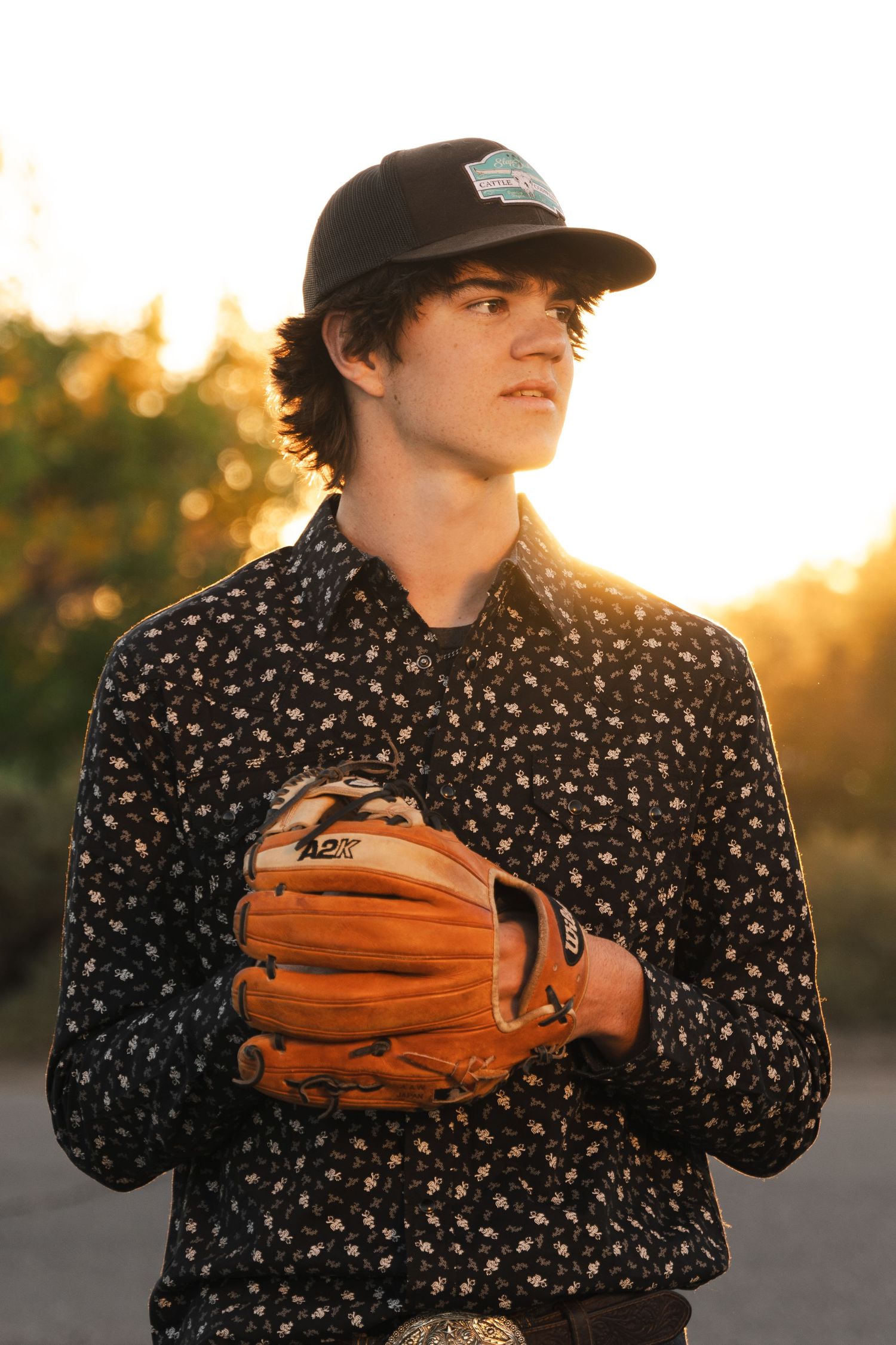 Baseball player wearing a patterned shirt holds up a brown leather glove in sunset lighting.