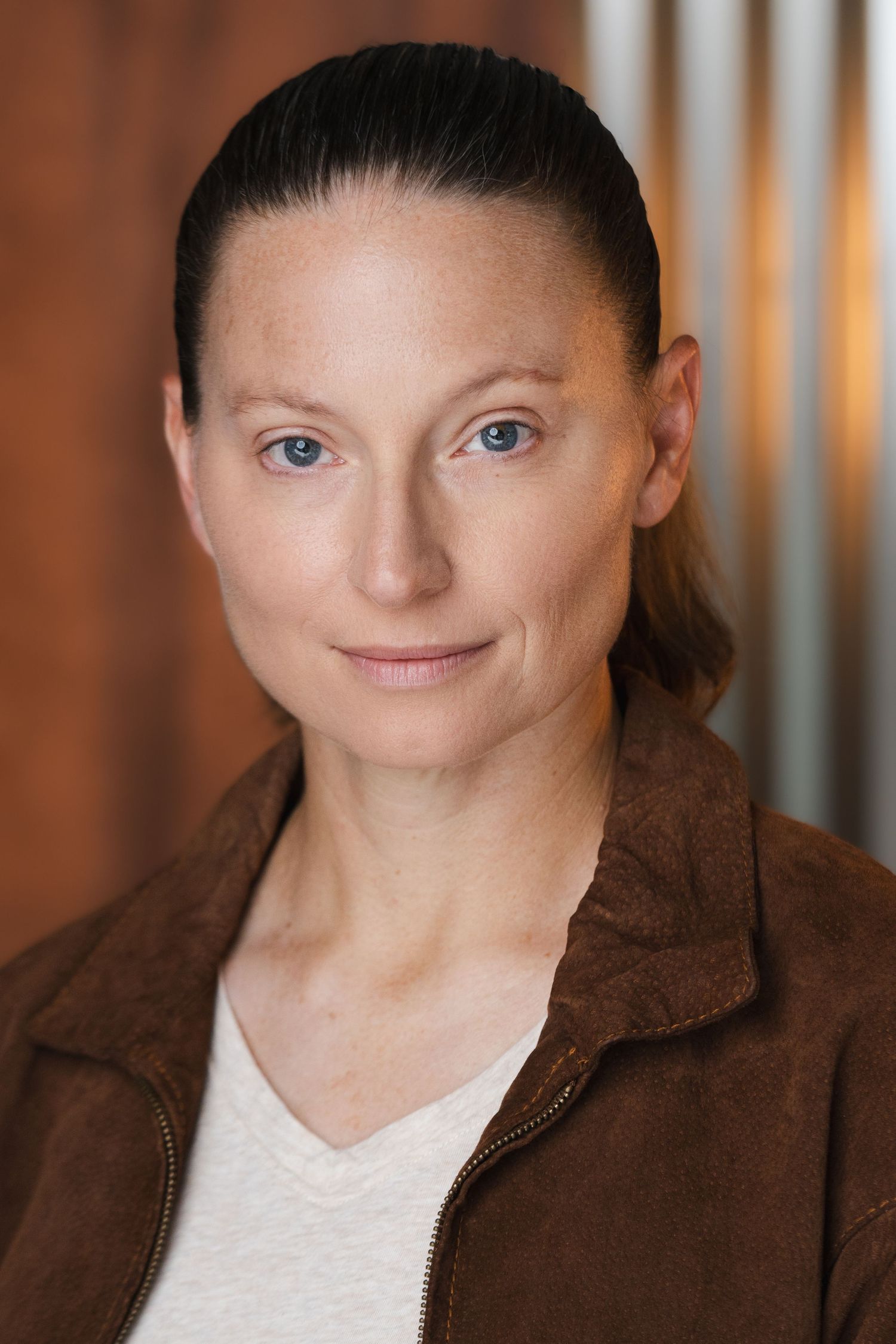 Professional headshot of a person with pulled-back hair wearing a brown jacket against a metallic background.