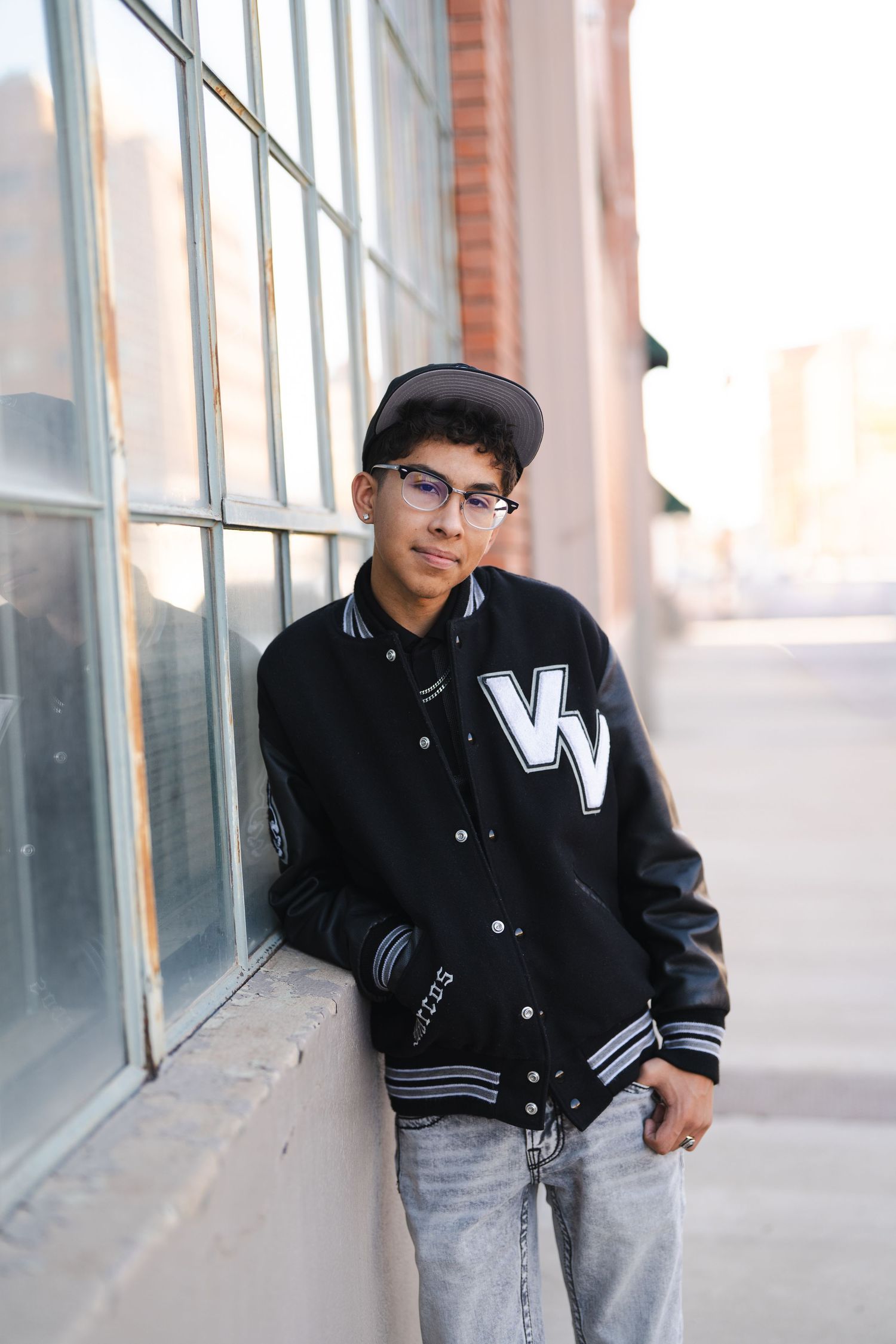 Person in a black and white varsity jacket and jeans leaning against a building with windows.