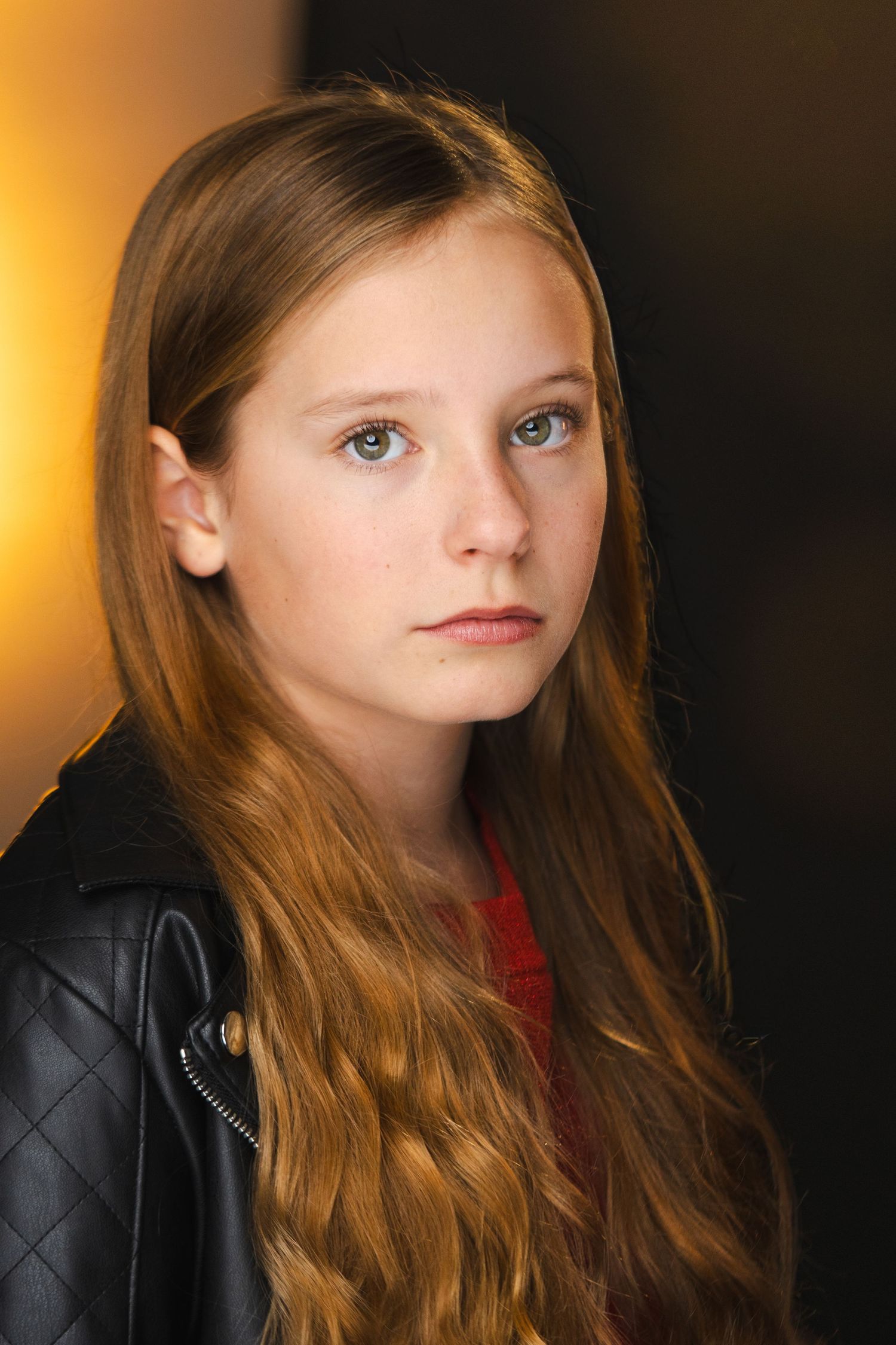 Portrait of a young person with long auburn hair wearing a black leather jacket against a dark background.