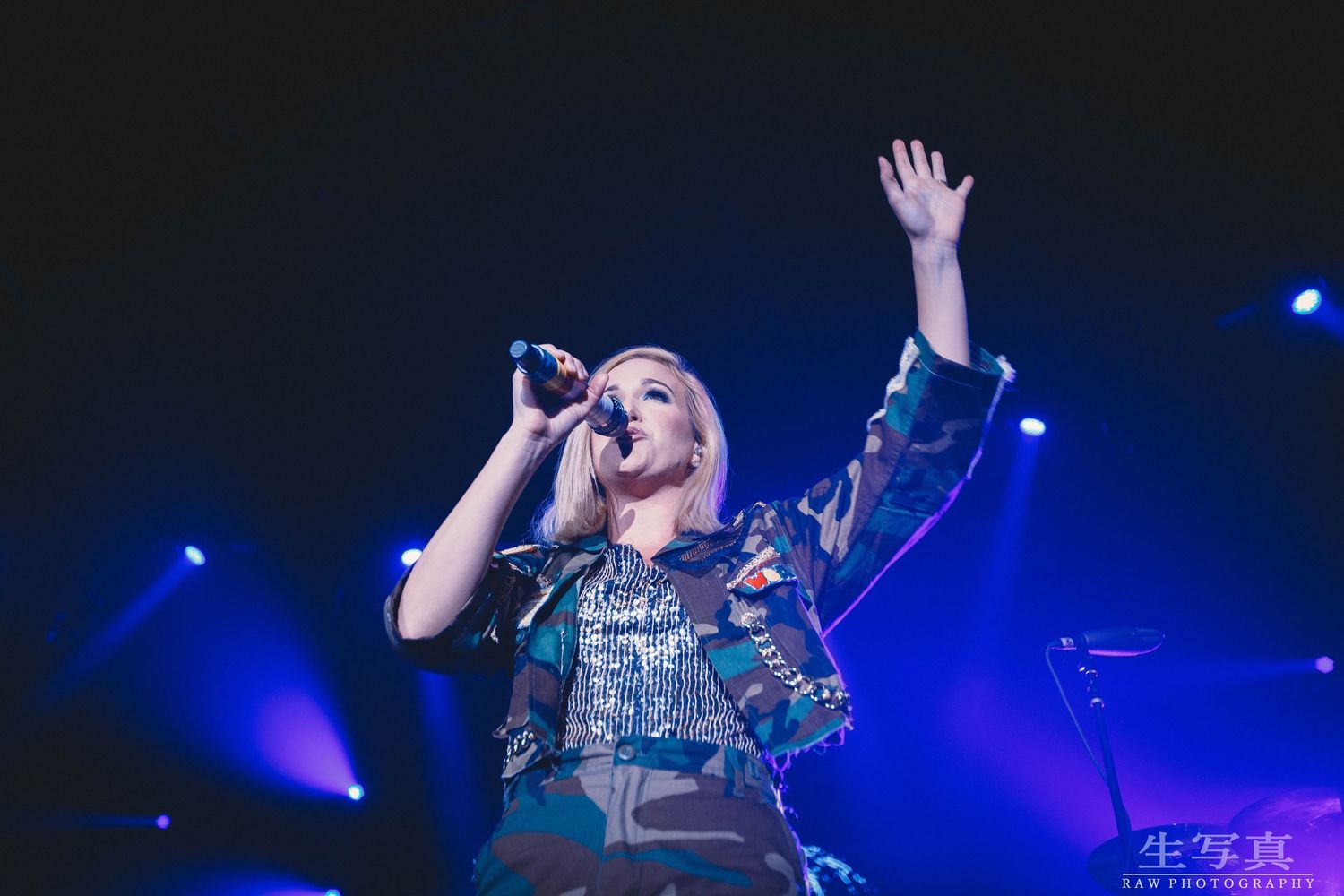 Performer singing into microphone with raised arm during energetic concert performance with blue stage lighting.