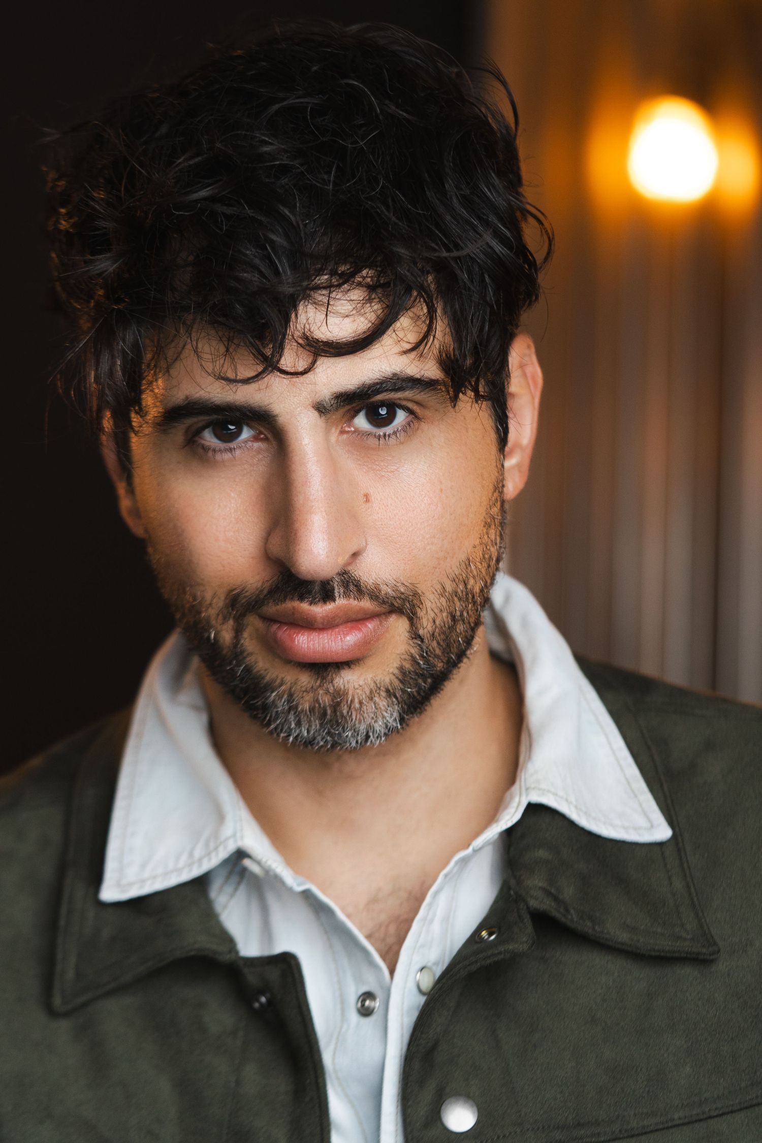 Professional headshot of person in olive jacket and white shirt against warm lighting background.