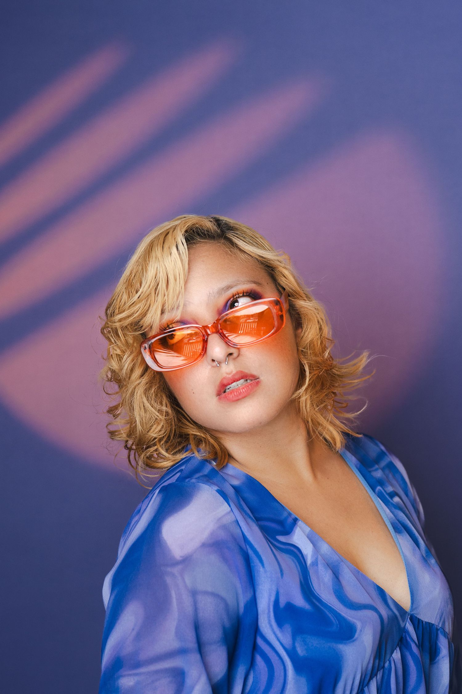Fashion portrait wearing orange tinted sunglasses and blue top against purple background.