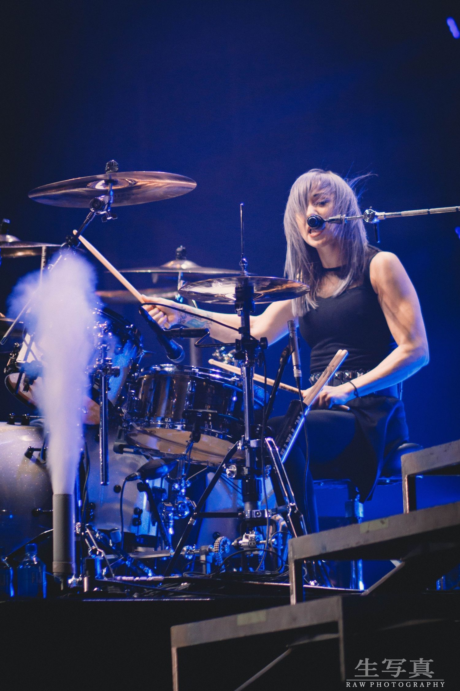Drummer performing energetically on stage with smoke effects and blue lighting.
