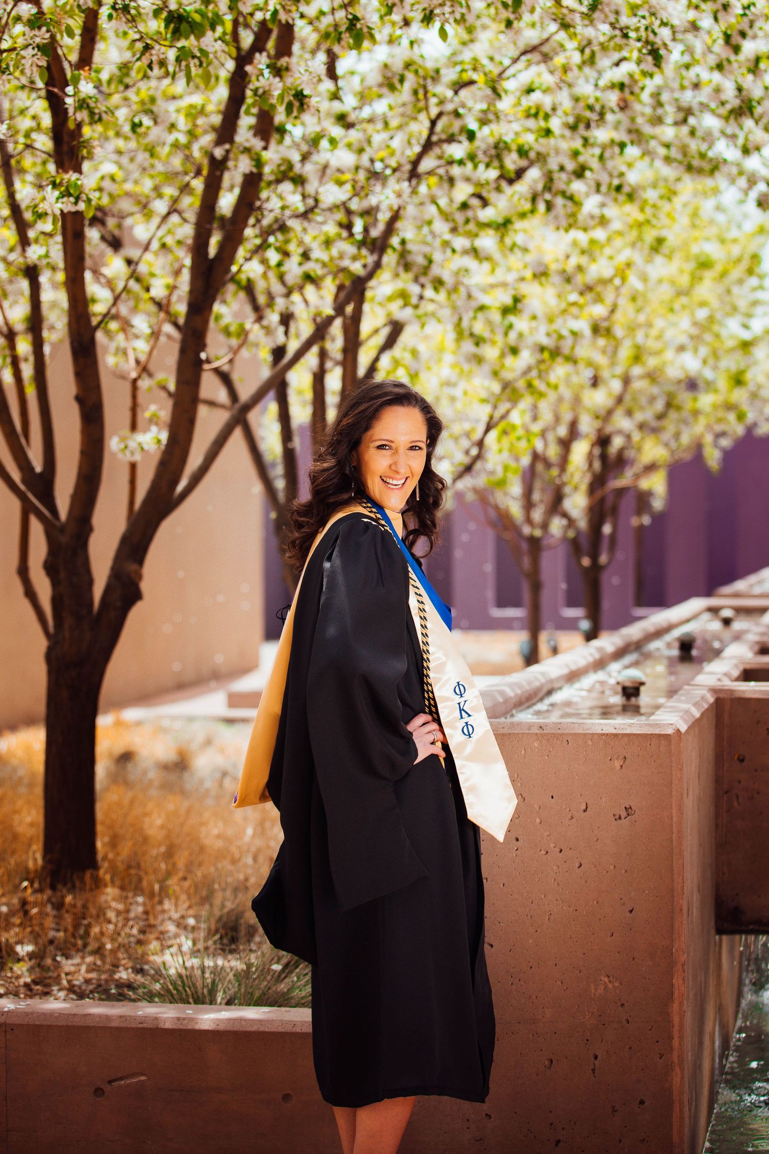 New Mexico Photographer - Graduate in black cap and gown poses outdoors on campus with blooming trees and brick walls in the background.