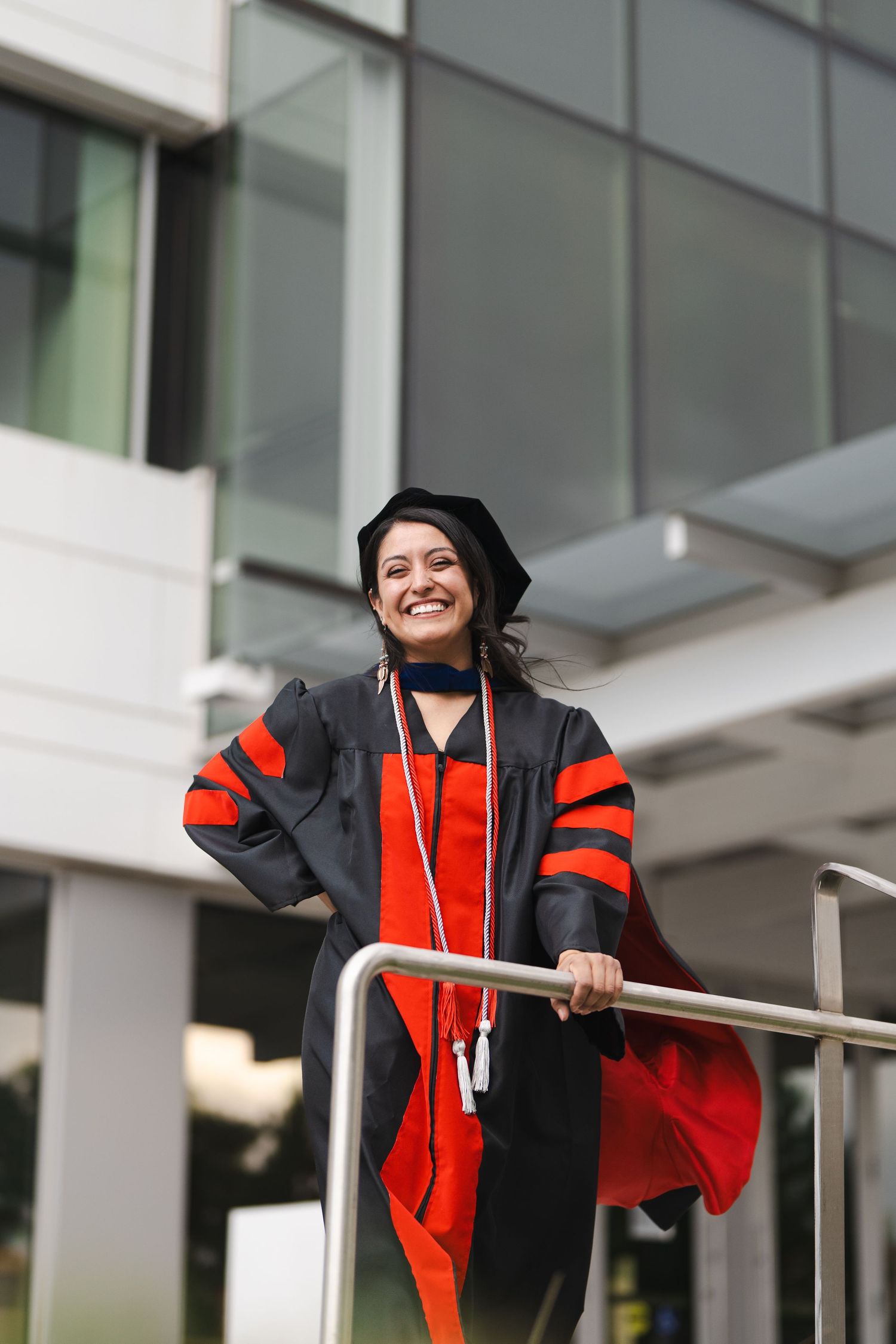 Graduate in academic regalia poses joyfully on campus building balcony.