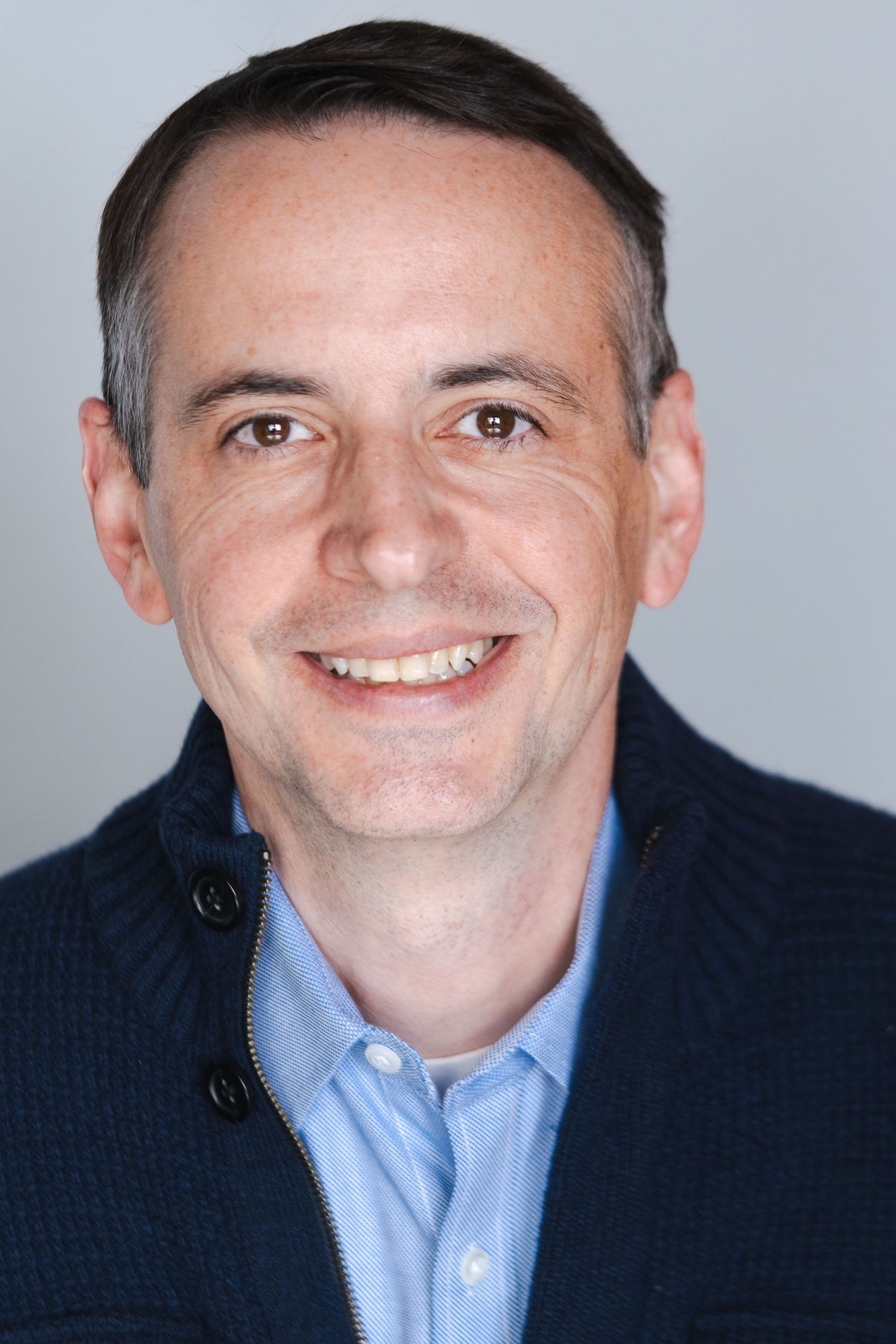 Business headshot of person in navy jacket and light blue shirt against gray background.