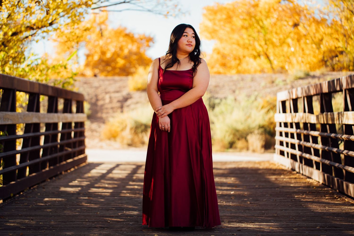 A woman in a burgundy dress poses on a wooden bridge during golden autumn.