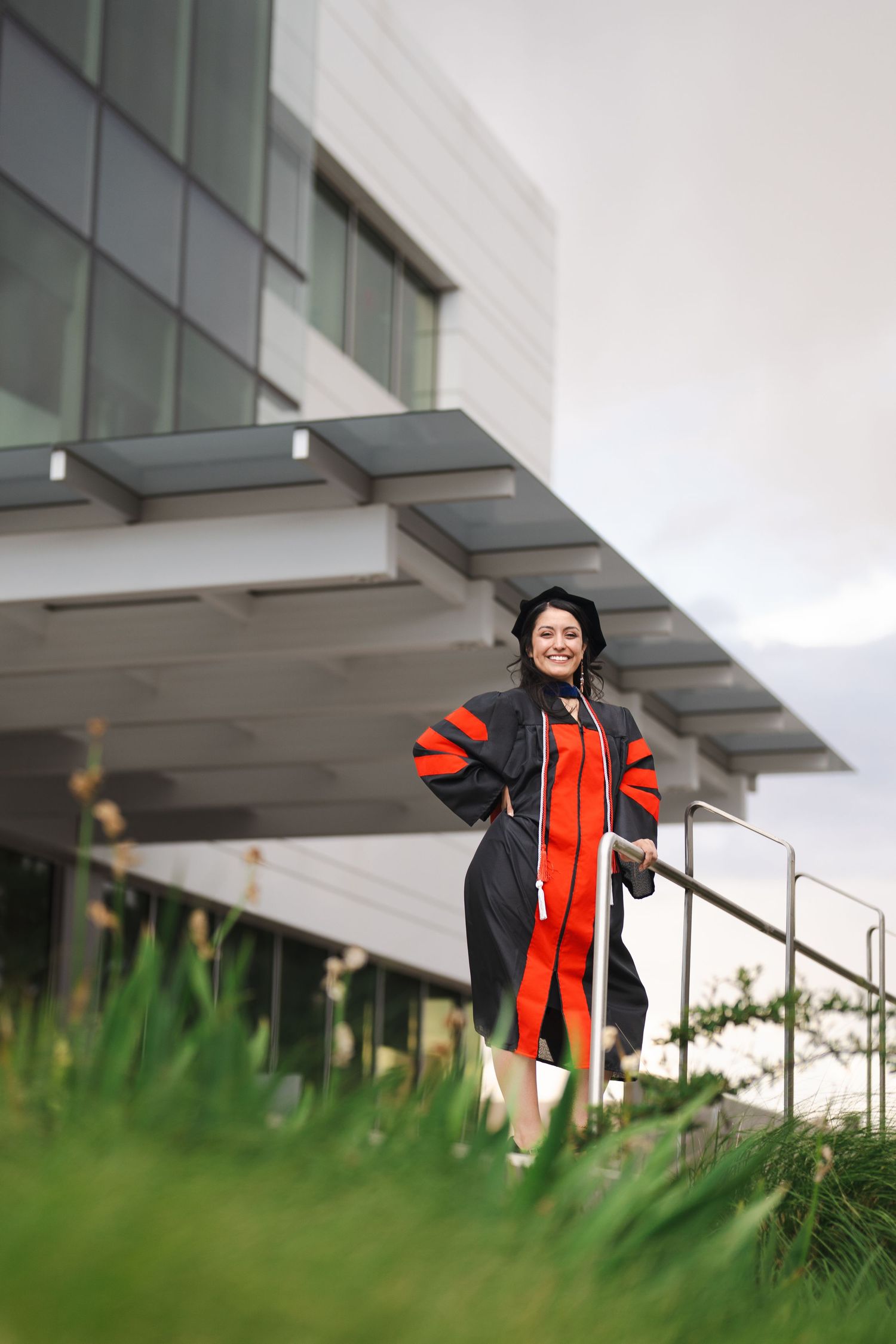 Graduate in red and black academic regalia photographed through foreground foliage.