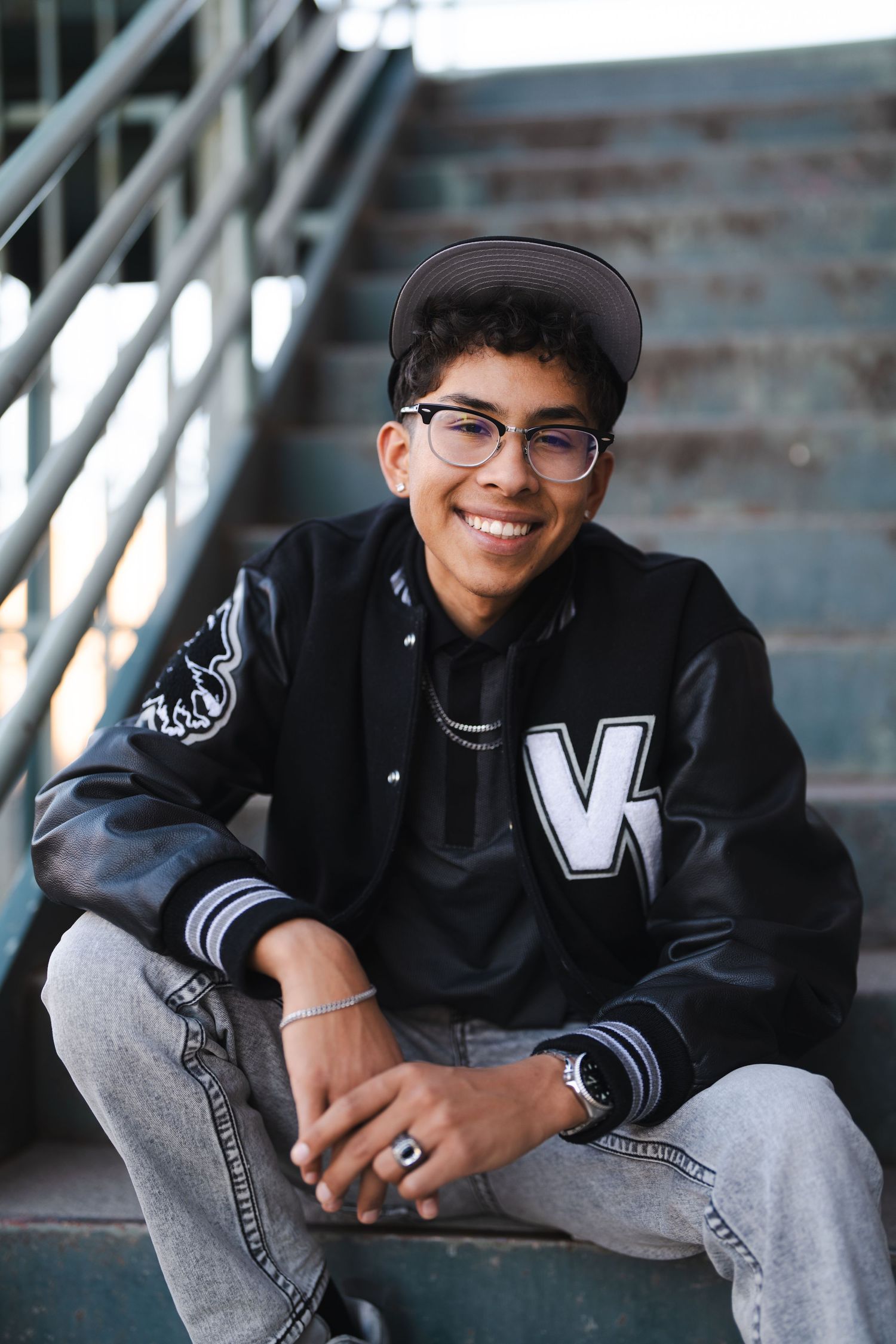 Student sitting on concrete steps wearing varsity jacket and glasses.