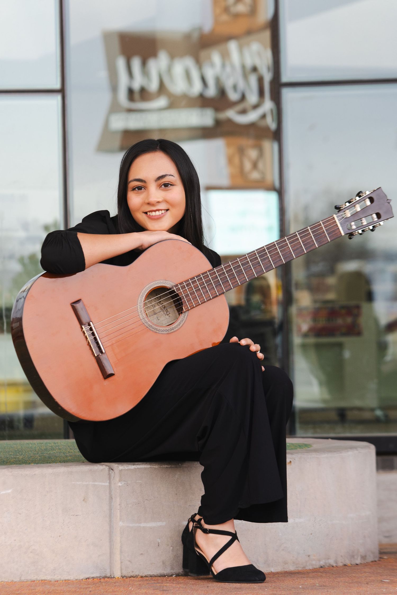Classical guitar musician in black attire poses with acoustic instrument against modern building exterior.