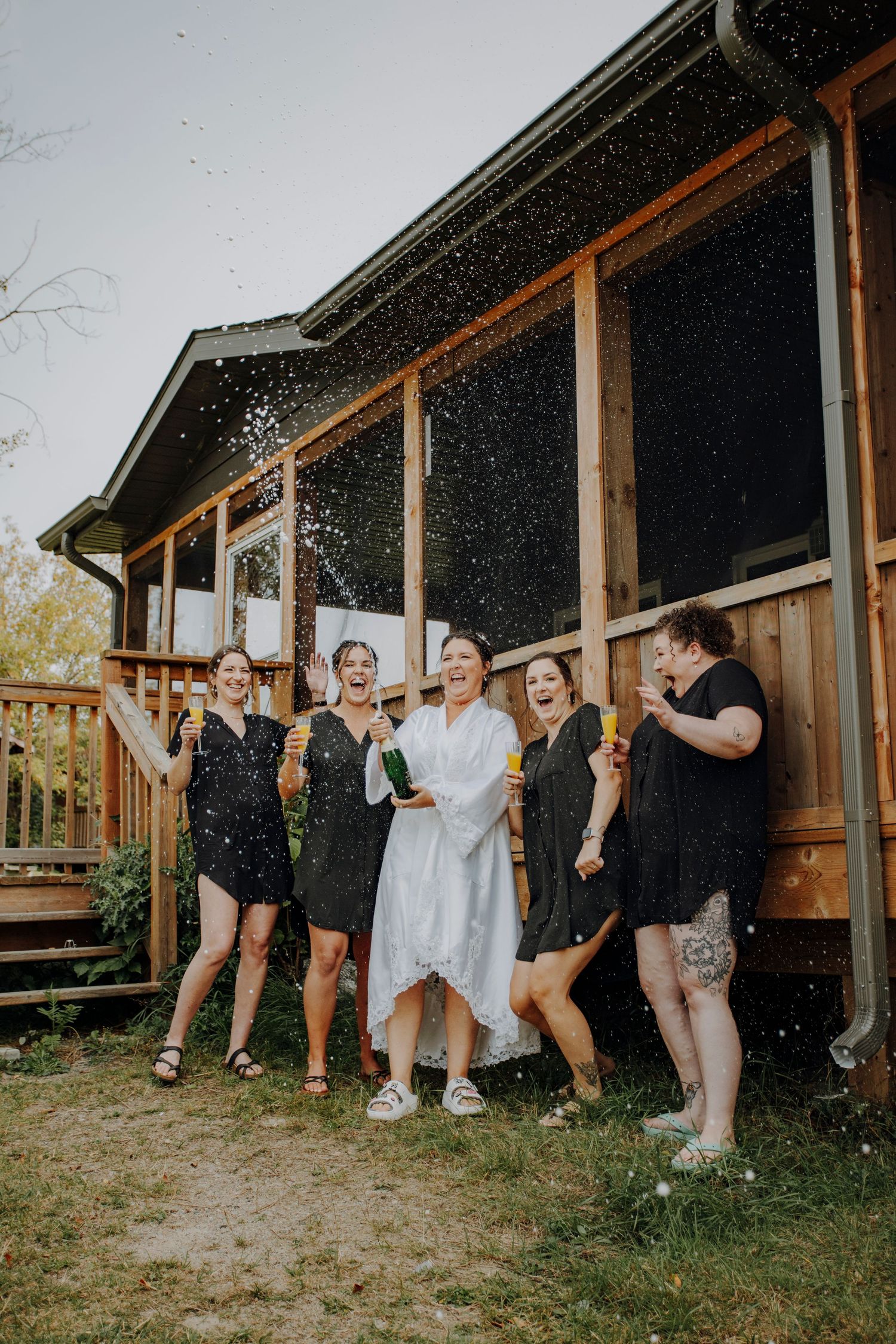 Wedding party in robes celebrating and spraying champagne outside rustic building.