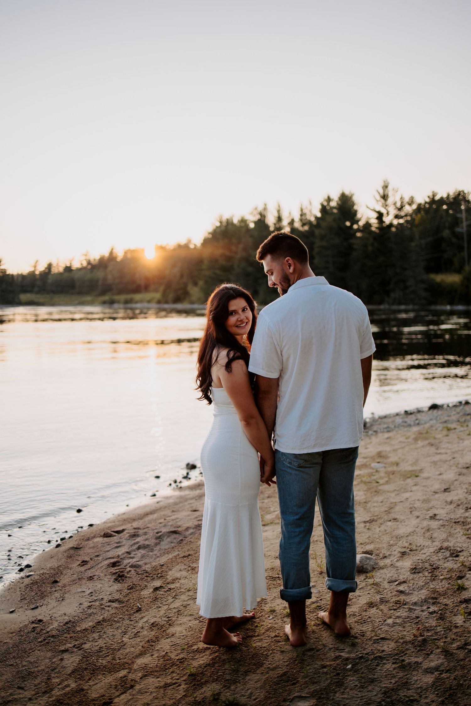Romantic sunset photo of a couple standing on a beach looking over a serene lake.